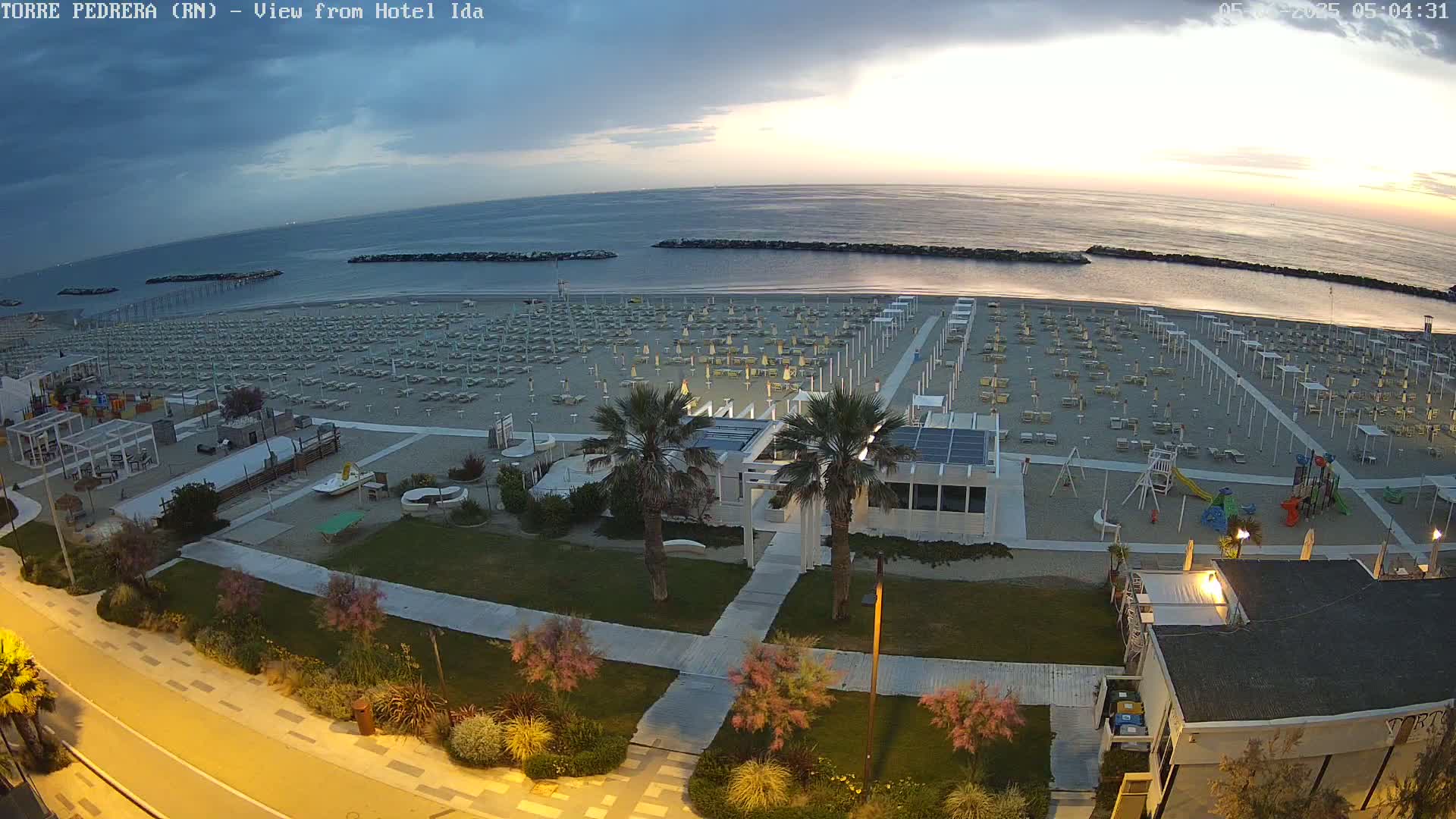An empty beach with rows of closed umbrellas and lounge chairs is seen at dawn under a mostly cloudy sky.