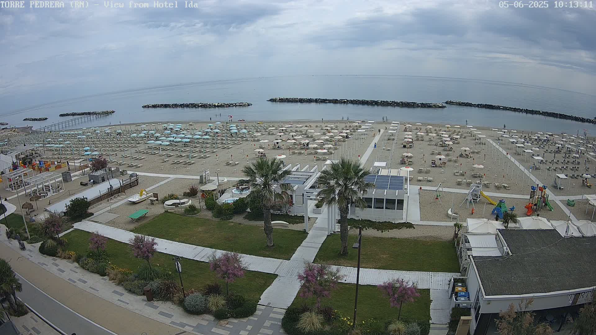 A beach with numerous umbrellas and lounge chairs under a partly cloudy sky, adjacent to a boardwalk, grassy area, and beachside buildings.