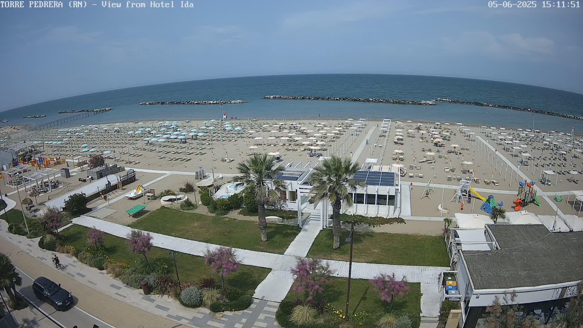 A beach with numerous umbrellas and lounge chairs, a playground, and a beachfront building is shown on a sunny day, next to a calm ocean.