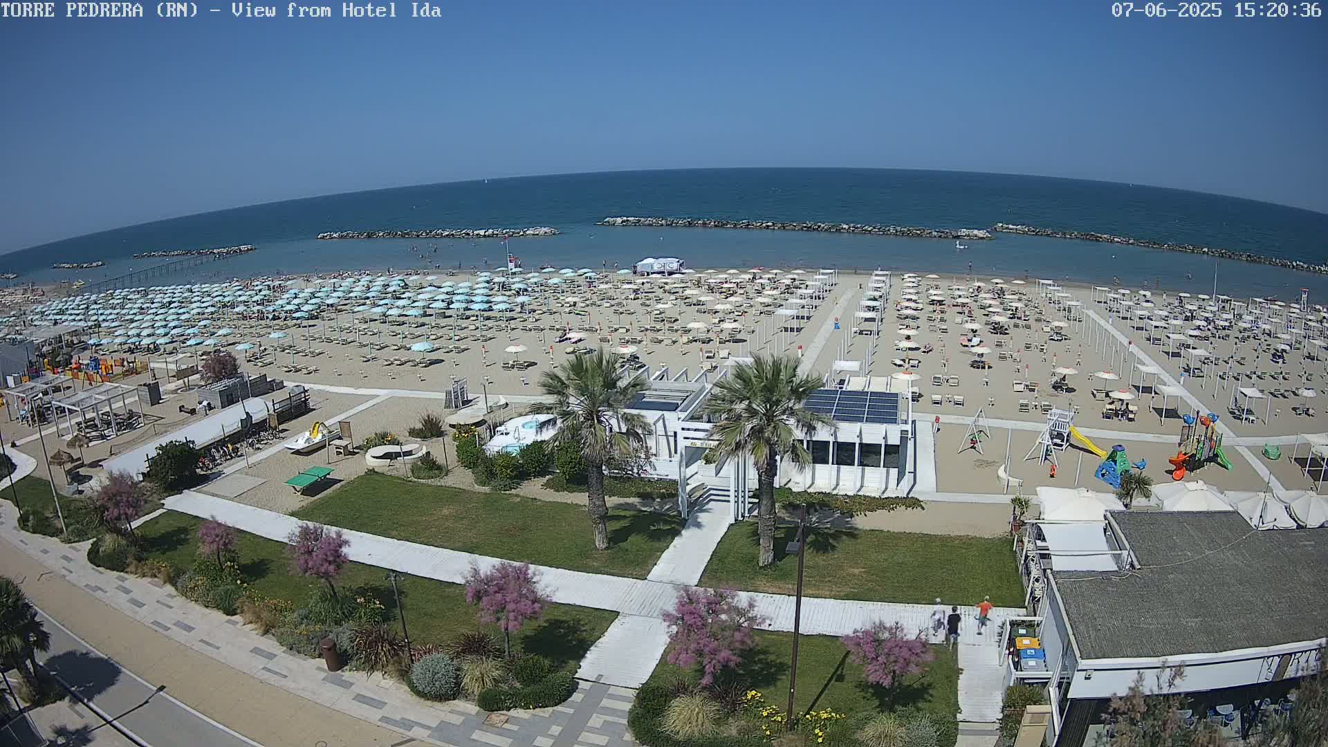 A sunny beach with numerous umbrellas and lounge chairs, a playground, and beachfront buildings is shown from an elevated viewpoint.