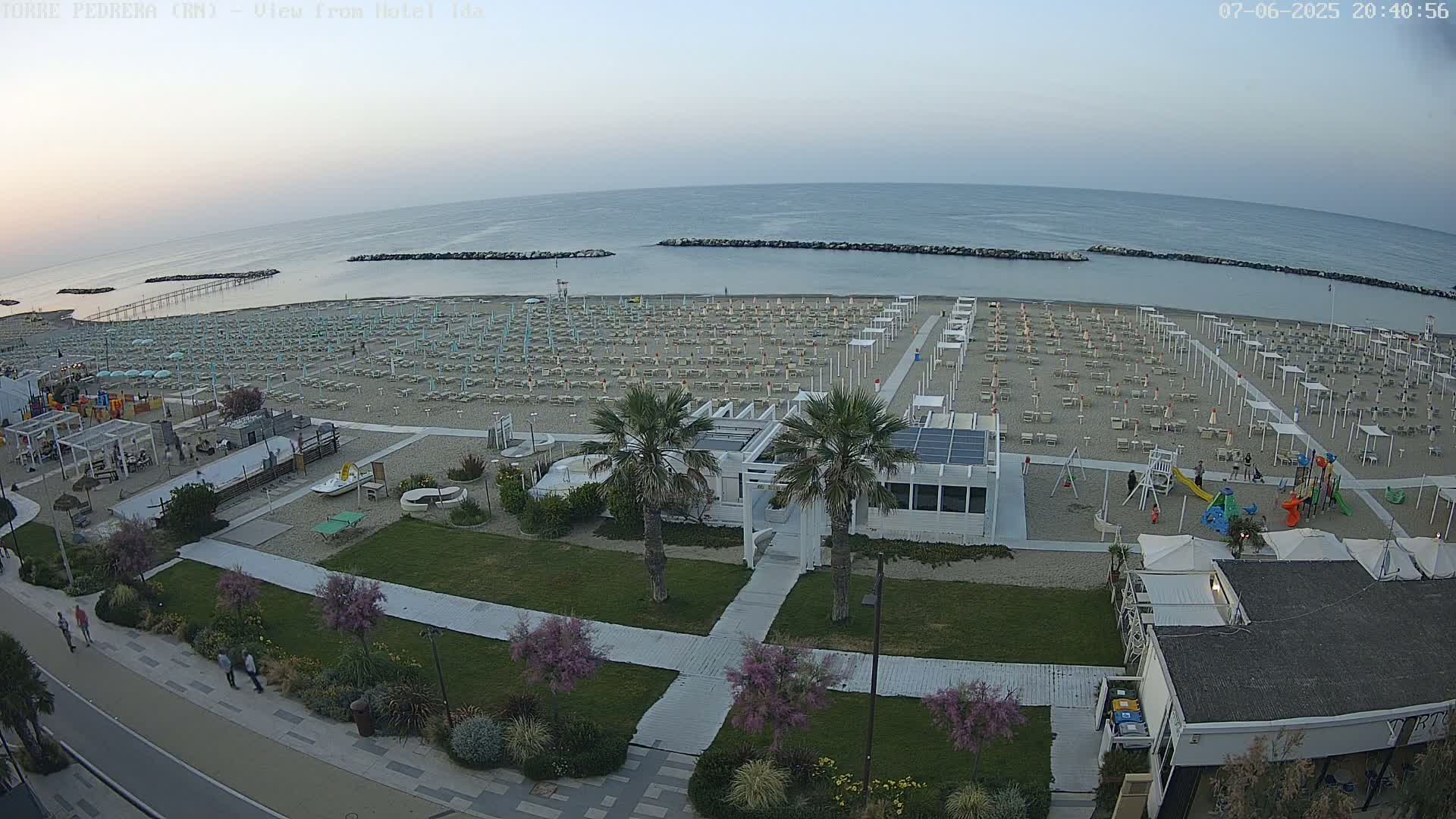 An aerial view of a beach at dusk shows rows of beach umbrellas and chairs, a playground, and a beachfront building, with calm water and a clear sky visible in the background.