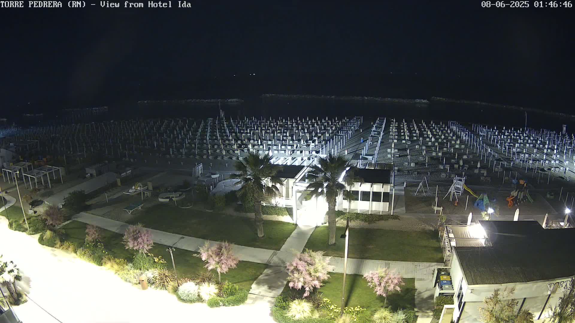 A nighttime view of a beach with numerous closed umbrellas and beach chairs, a playground, and some palm trees, all illuminated under dark skies.