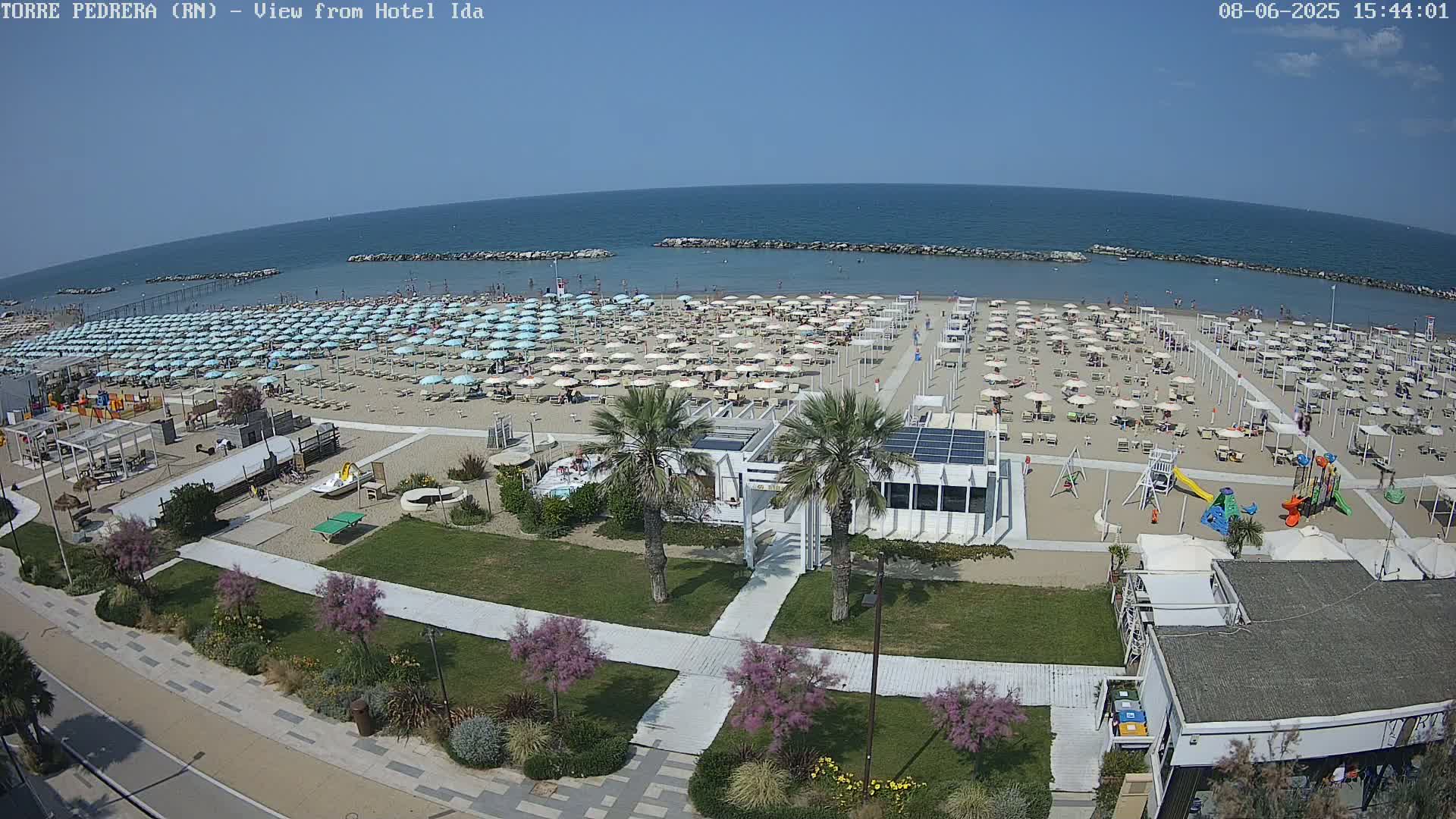 A sunny beach with numerous umbrellas, beachgoers, and a playground, situated next to a paved walkway and green landscaping, overlooks a calm sea.