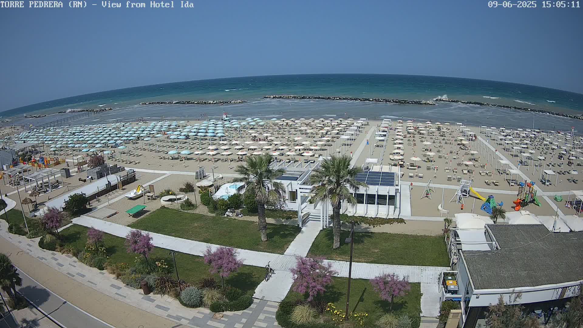 An aerial view of a beach on a sunny day shows numerous beach umbrellas and lounge chairs on the sand, with a playground, small buildings, and palm trees nearby.