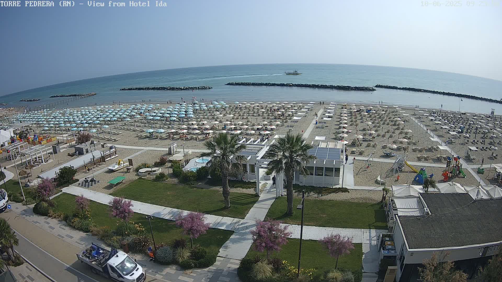 A sunny beach with numerous light-blue umbrellas, beachside amenities, and a playground, situated next to calm blue water.