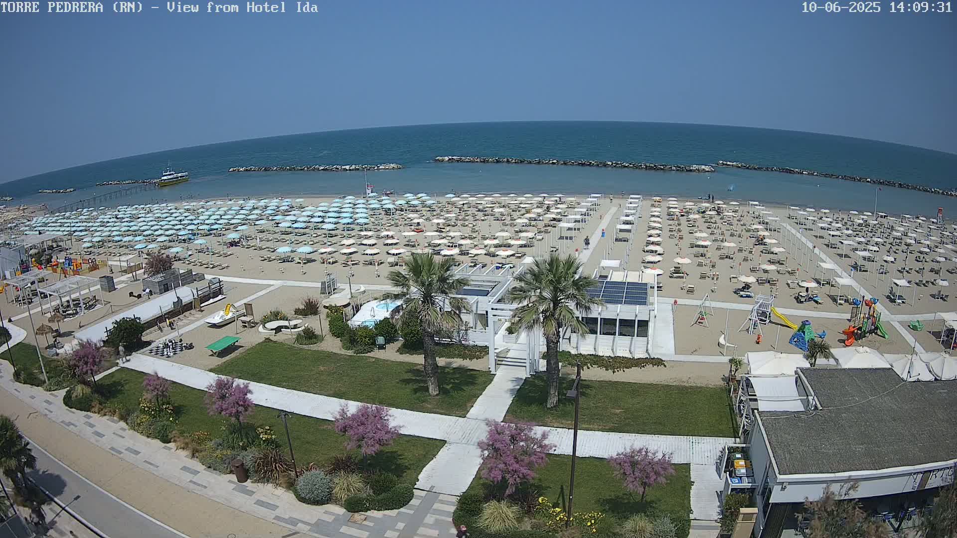A sandy beach with numerous light-blue umbrellas and lounge chairs under a clear blue sky, featuring a beachfront building and playground, is viewed from above.