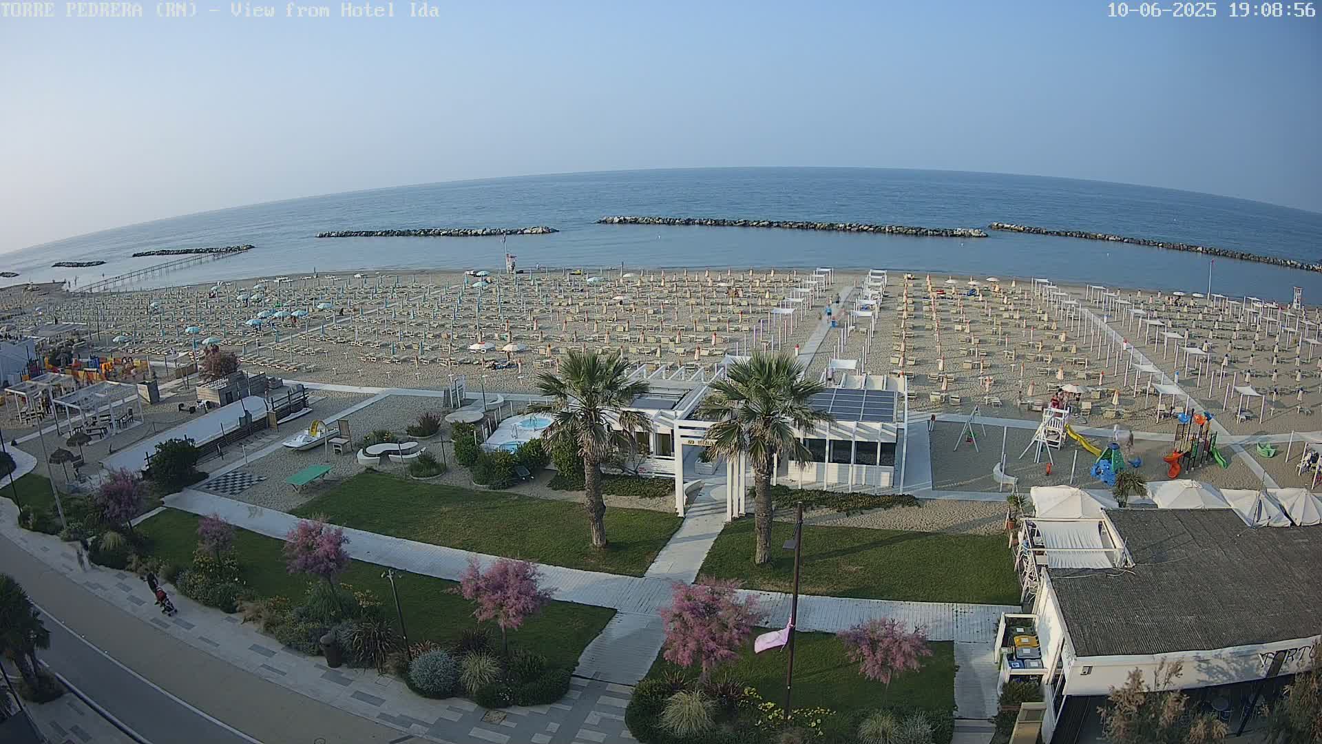 An aerial view of a beach with numerous umbrellas and lounge chairs, a playground, and palm trees, under a clear sky.