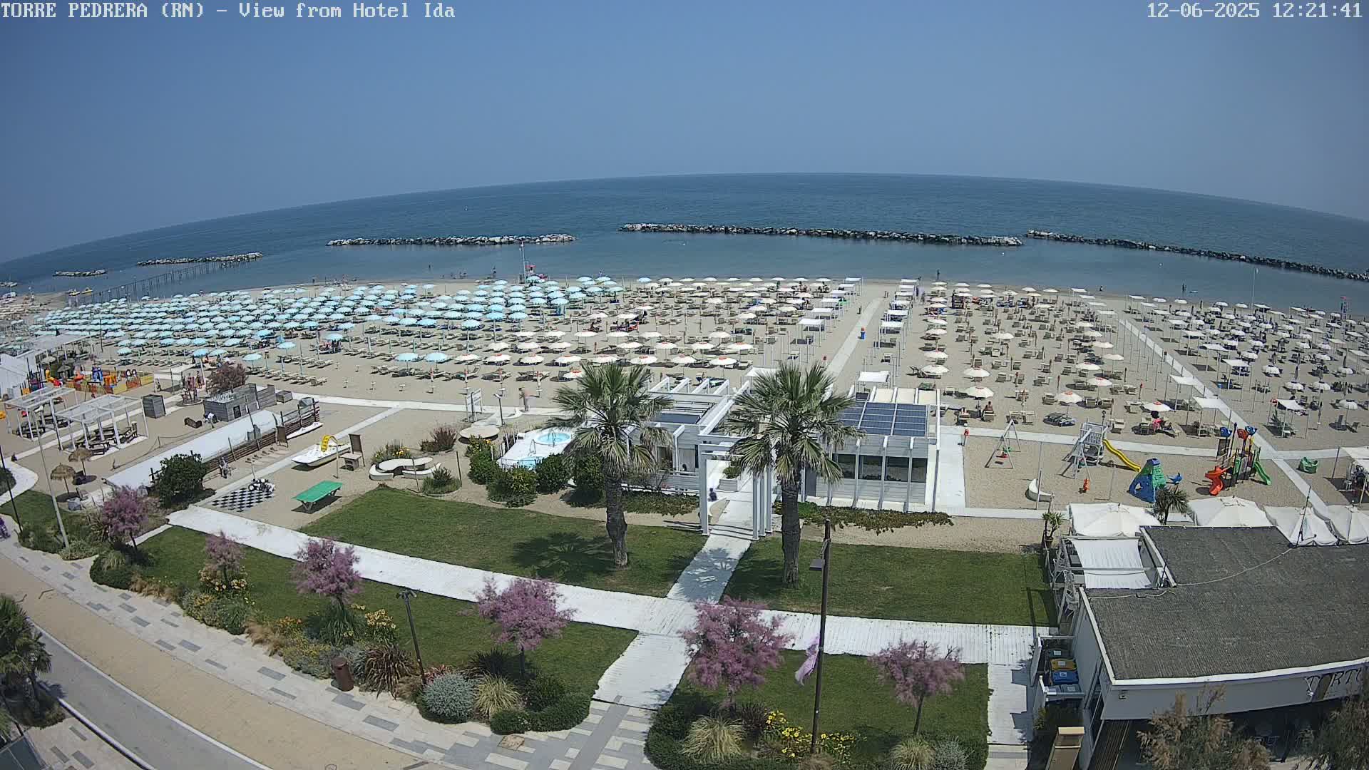 A sunny beach scene shows a sandy beach filled with numerous light-blue umbrellas, beach chairs, and a children's playground adjacent to a beachfront building with palm trees and a walkway.