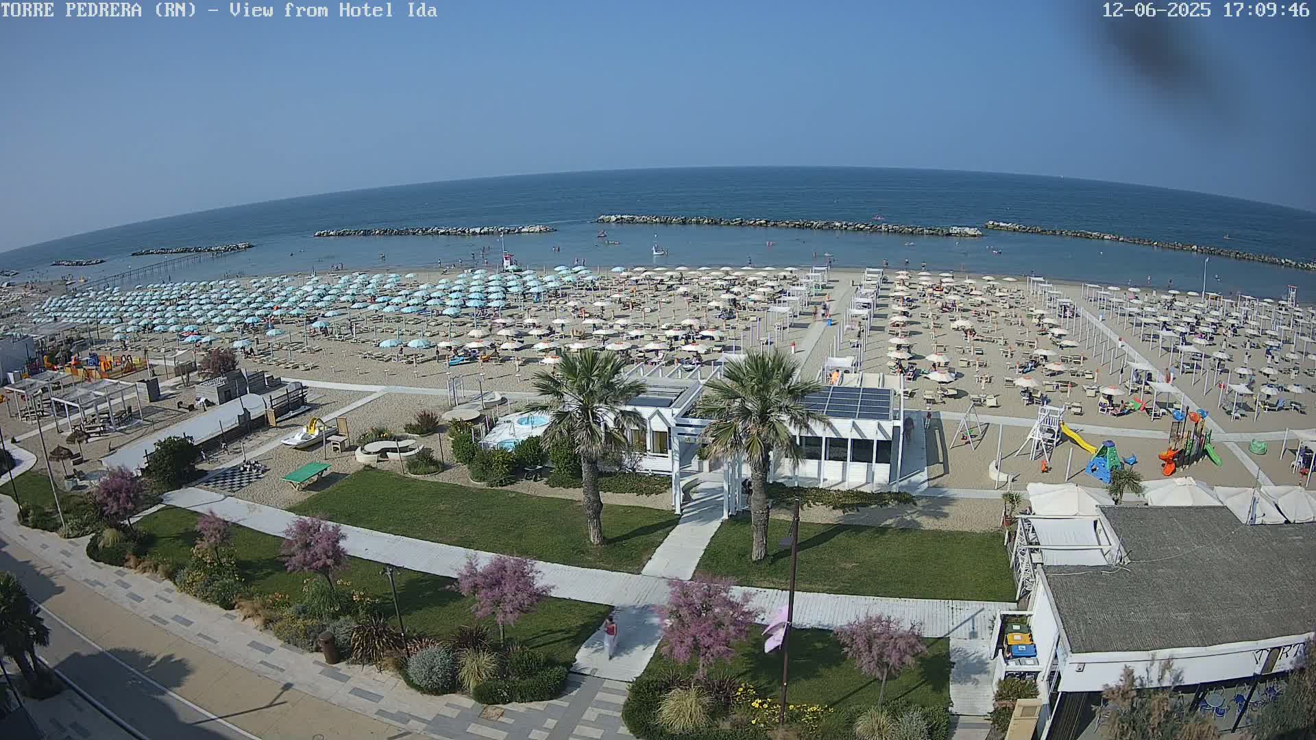 A sunny beach scene shows numerous light-blue umbrellas and beachgoers on a sandy shore, with a calm ocean beyond and a grassy area with palm trees and a building closer to the viewer.