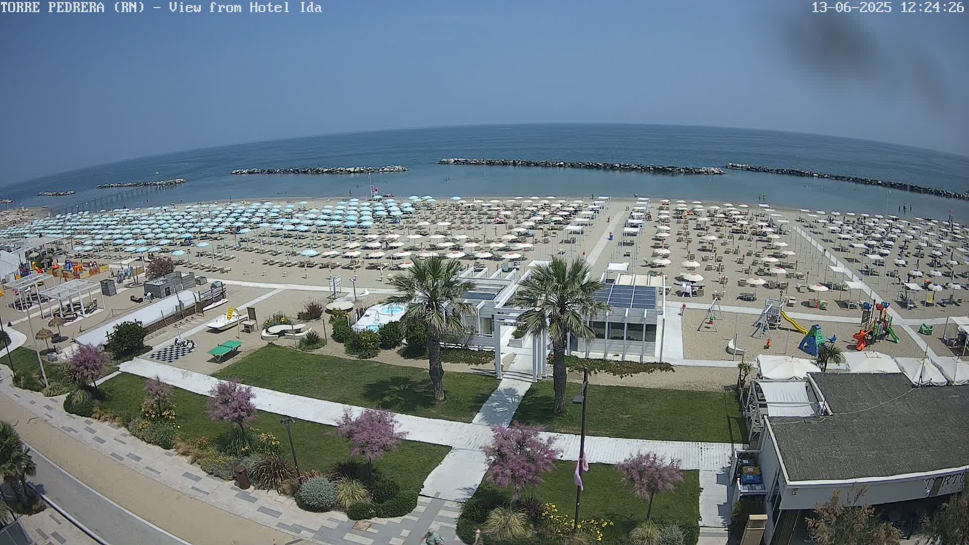 A sunny day at a beach shows numerous beach umbrellas and lounge chairs on the sand, with a playground and other beach amenities nearby.