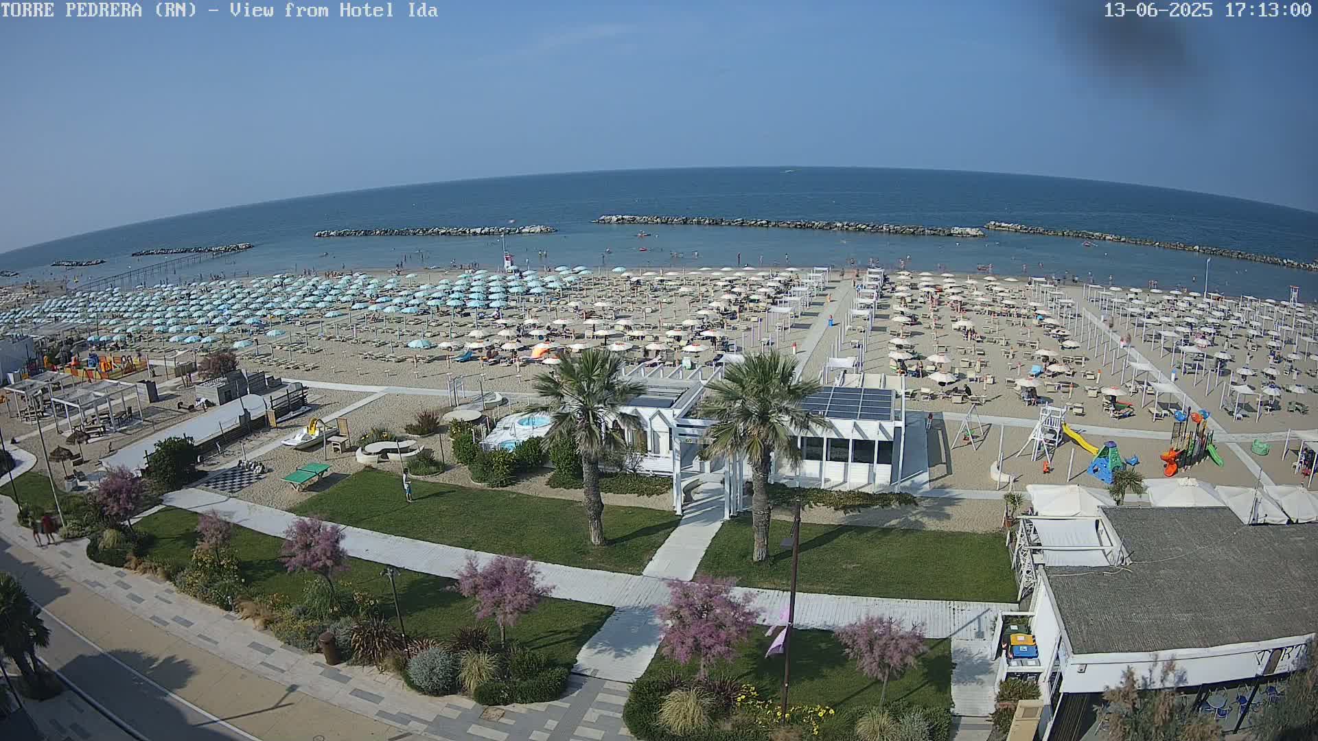 A beach scene on a sunny day shows numerous beach umbrellas and lounge chairs set up on a sandy beach, with a calm sea in the background, all overseen from an elevated position.