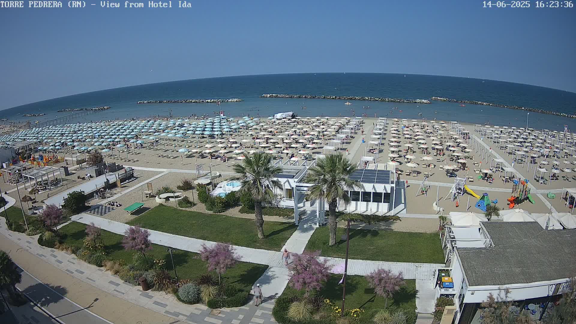 A sunny beach scene shows a sandy shoreline densely populated with umbrellas and beachgoers, children's playground equipment, and beachfront buildings under a clear sky.