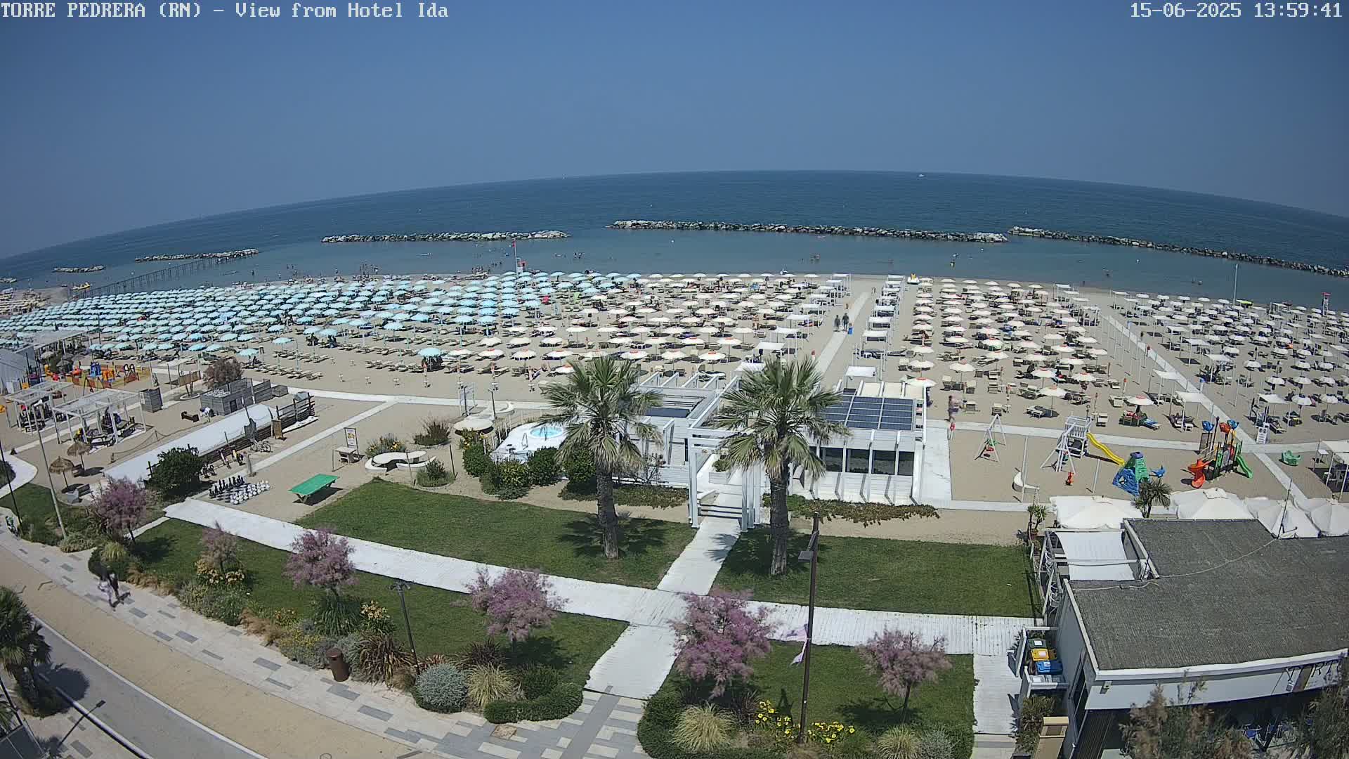 A sunny beach scene with numerous light blue umbrellas, a playground, and beachside buildings.