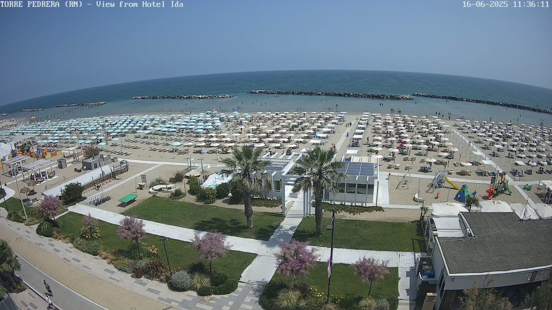 A sandy beach with numerous umbrellas and sunbathers is seen from above on a sunny day, next to a grassy area with pathways and a few buildings.