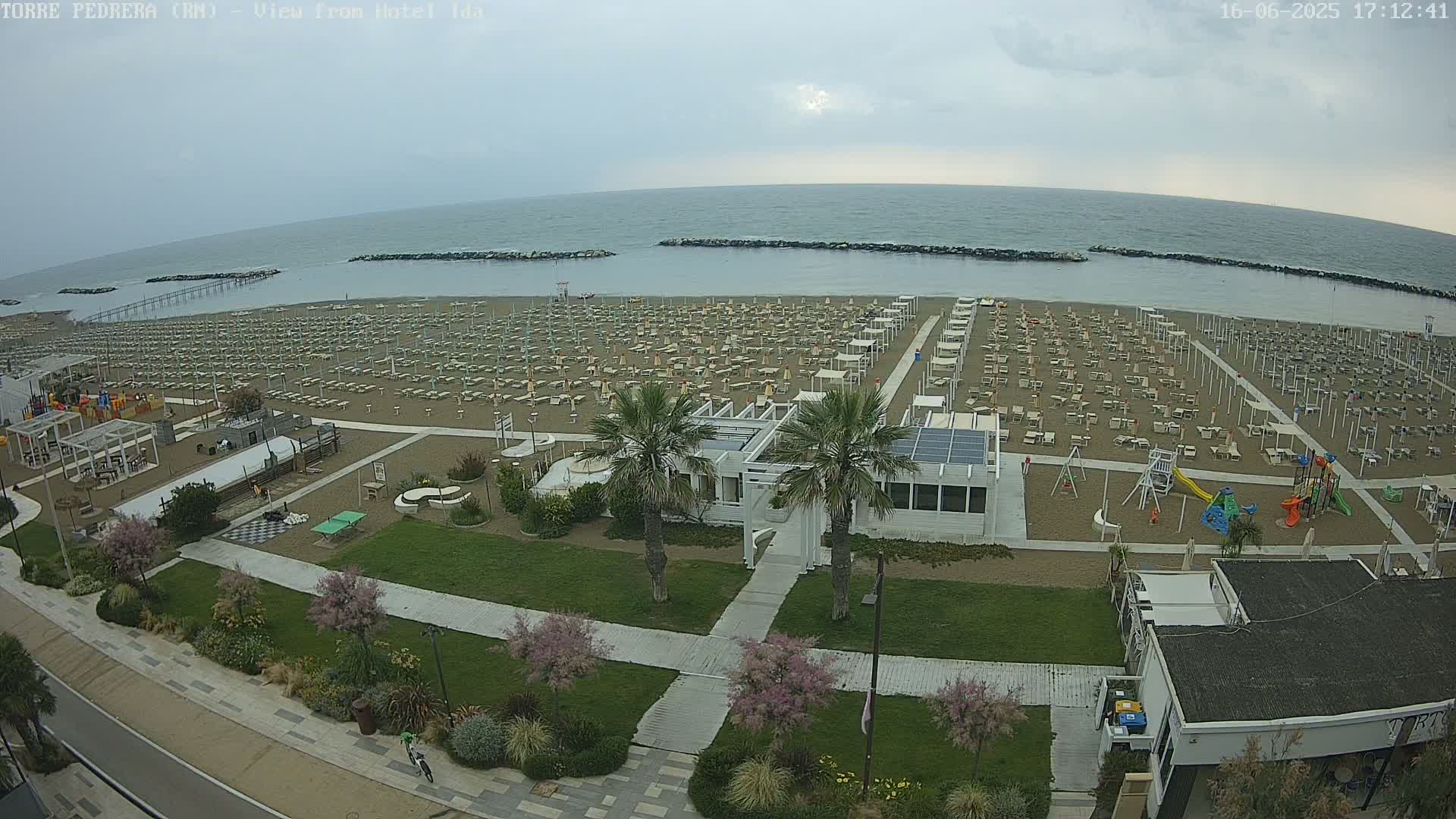 An aerial view shows a beach with numerous beach umbrellas and lounge chairs under an overcast sky, adjacent to a grassy area with landscaping, walkways, and a building.
