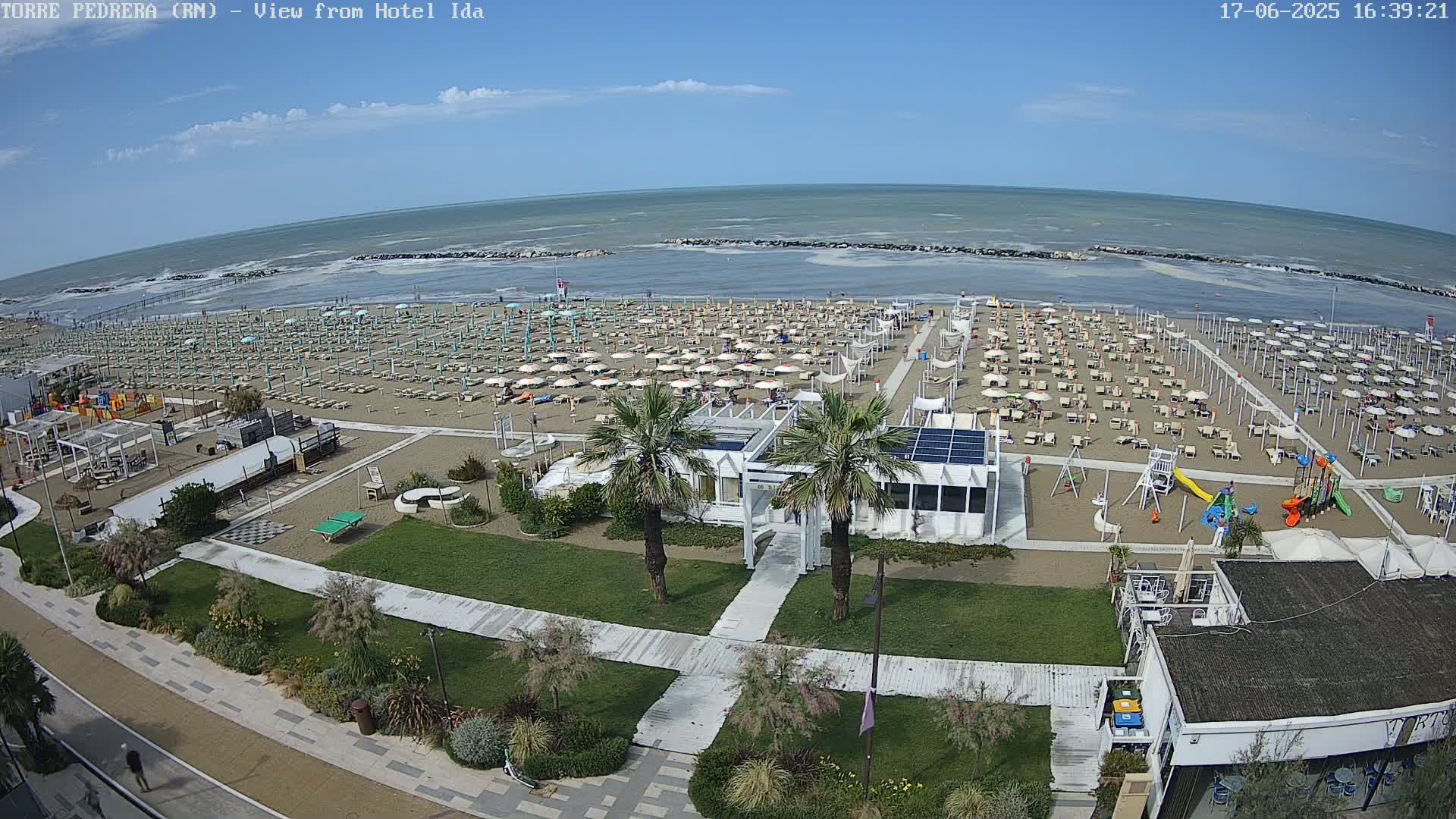 A sandy beach with numerous umbrellas and lounge chairs, a playground, and a beachfront building is visible under a sunny sky, with a moderately wavy ocean in the background.