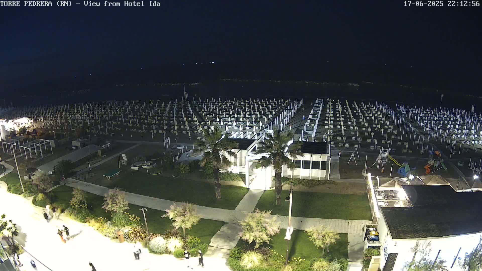 A nighttime aerial view of a beach with rows of closed beach umbrellas, a playground, and a few people walking along a paved path.