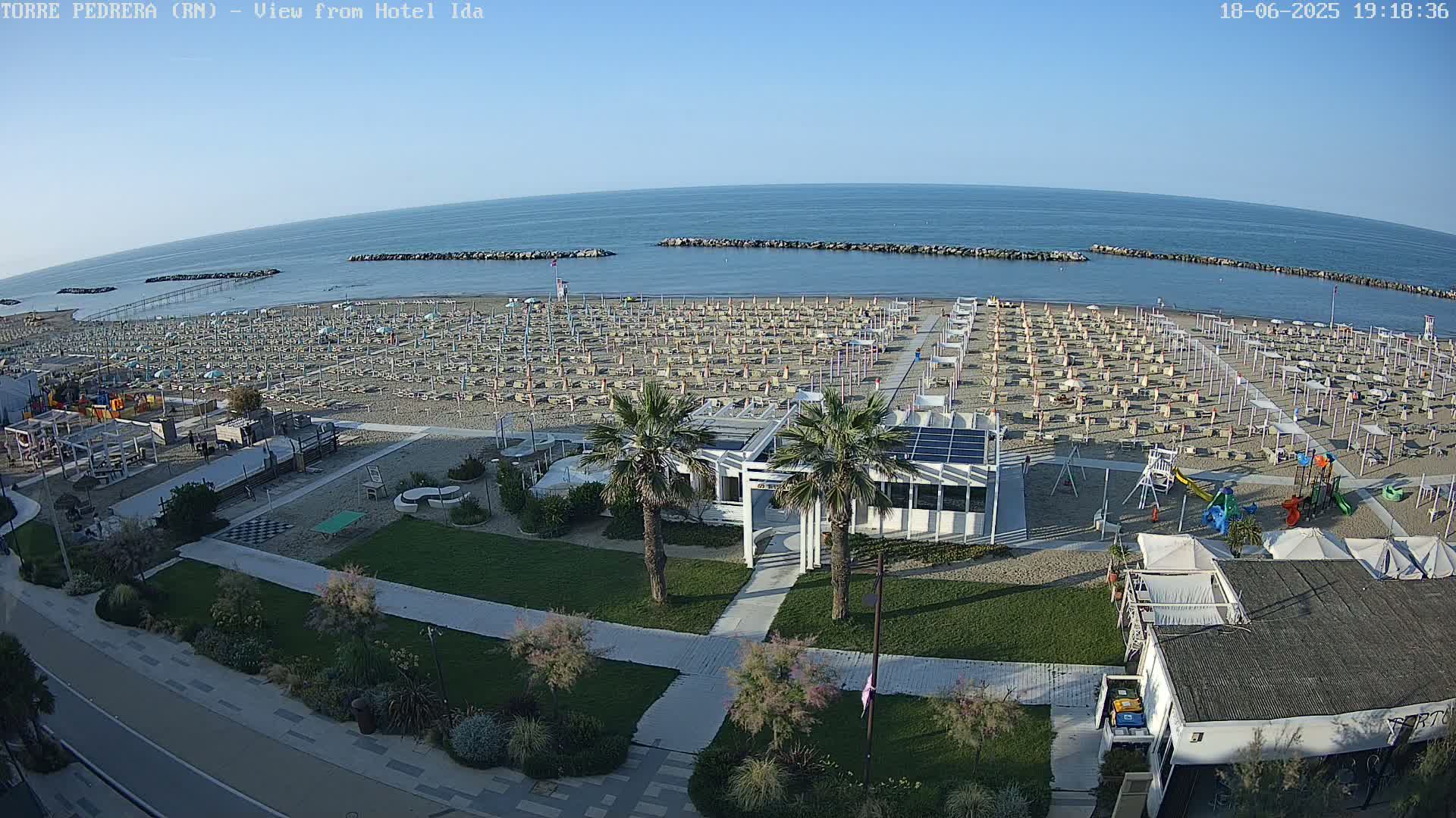 An expansive beach with numerous umbrellas and lounge chairs is visible, next to a calm ocean under a clear sky.