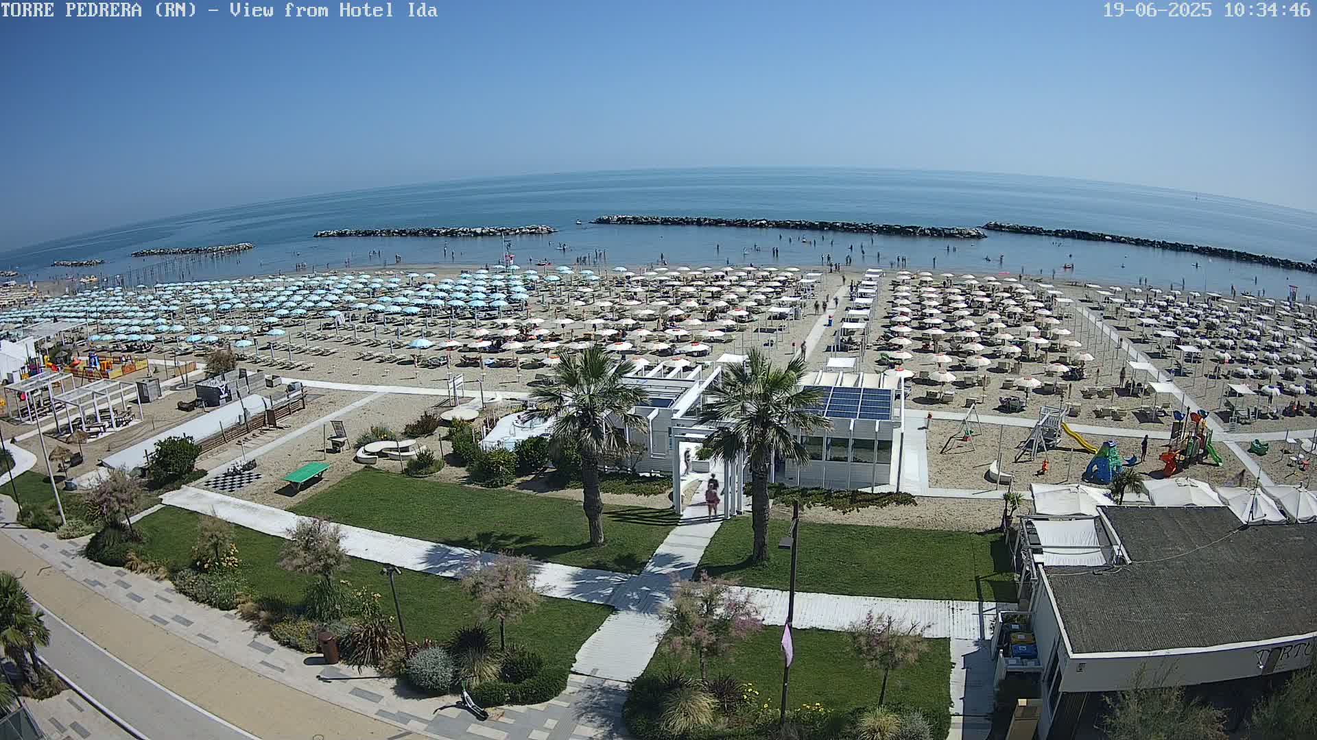 A sandy beach filled with numerous umbrellas and sunbathers is visible from an elevated viewpoint, under sunny skies, with calm ocean water in the background.