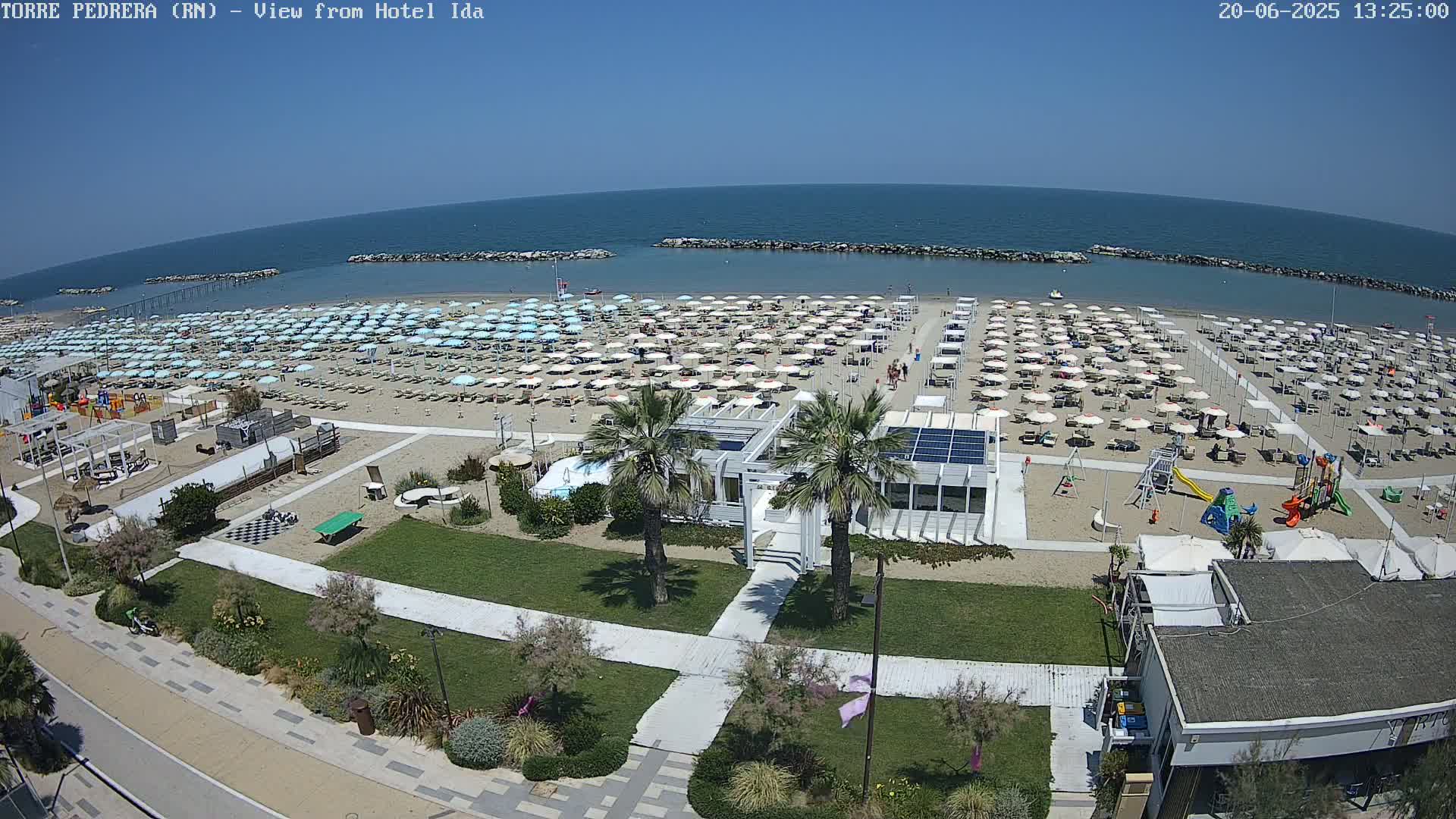 A sandy beach with numerous light-blue and white umbrellas, a playground, and beachside buildings is shown on a sunny day, next to a calm blue sea.