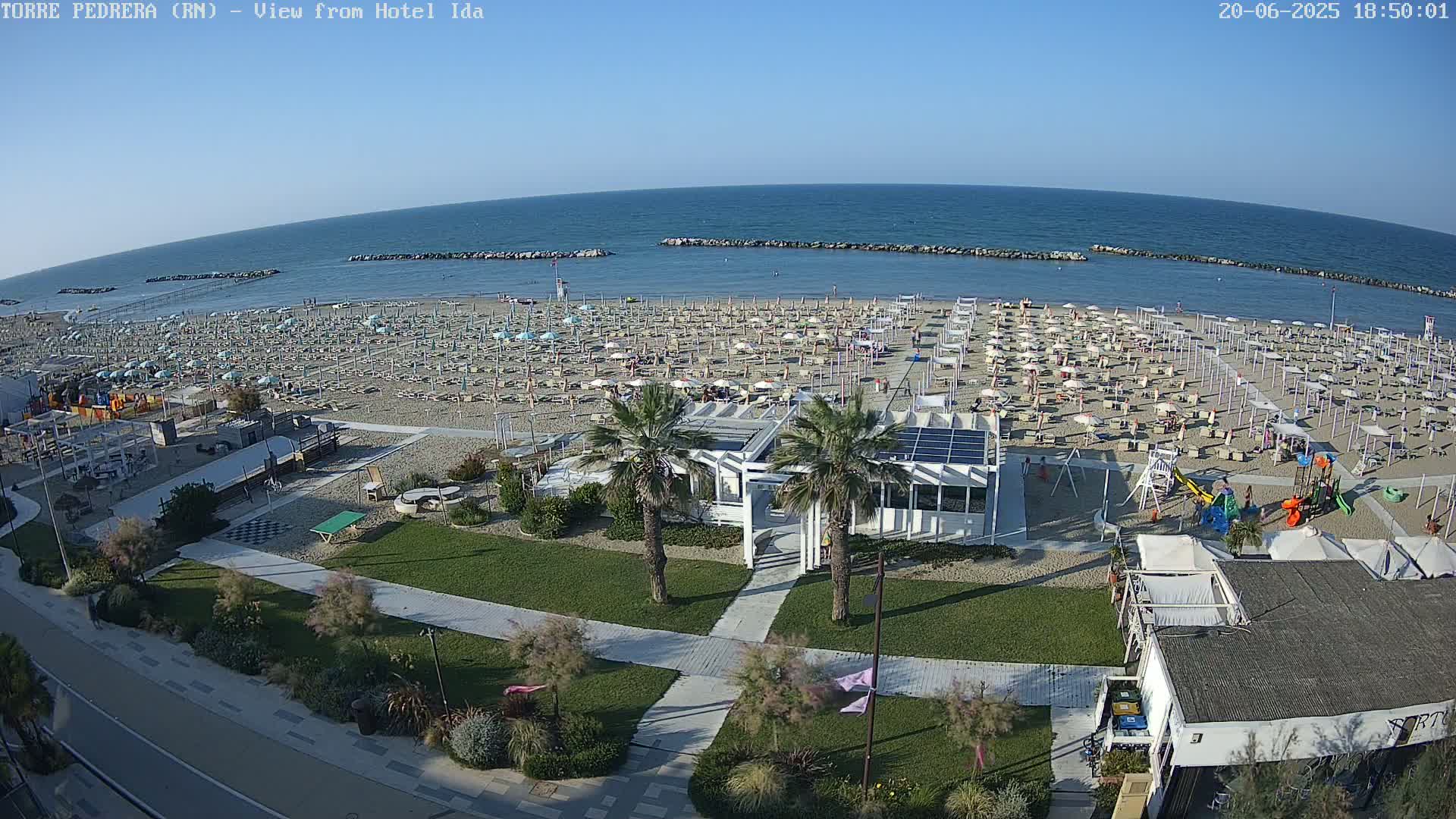 A beach with numerous umbrellas and lounge chairs is shown on a sunny day, adjacent to a grassy area with walkways, palm trees, and beachside buildings.