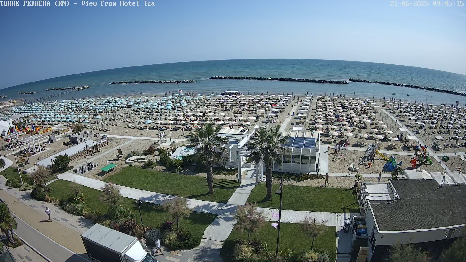 A sunny beach with numerous umbrellas and beachgoers, a playground, and beachside structures is visible from an elevated vantage point, overlooking a calm sea.
