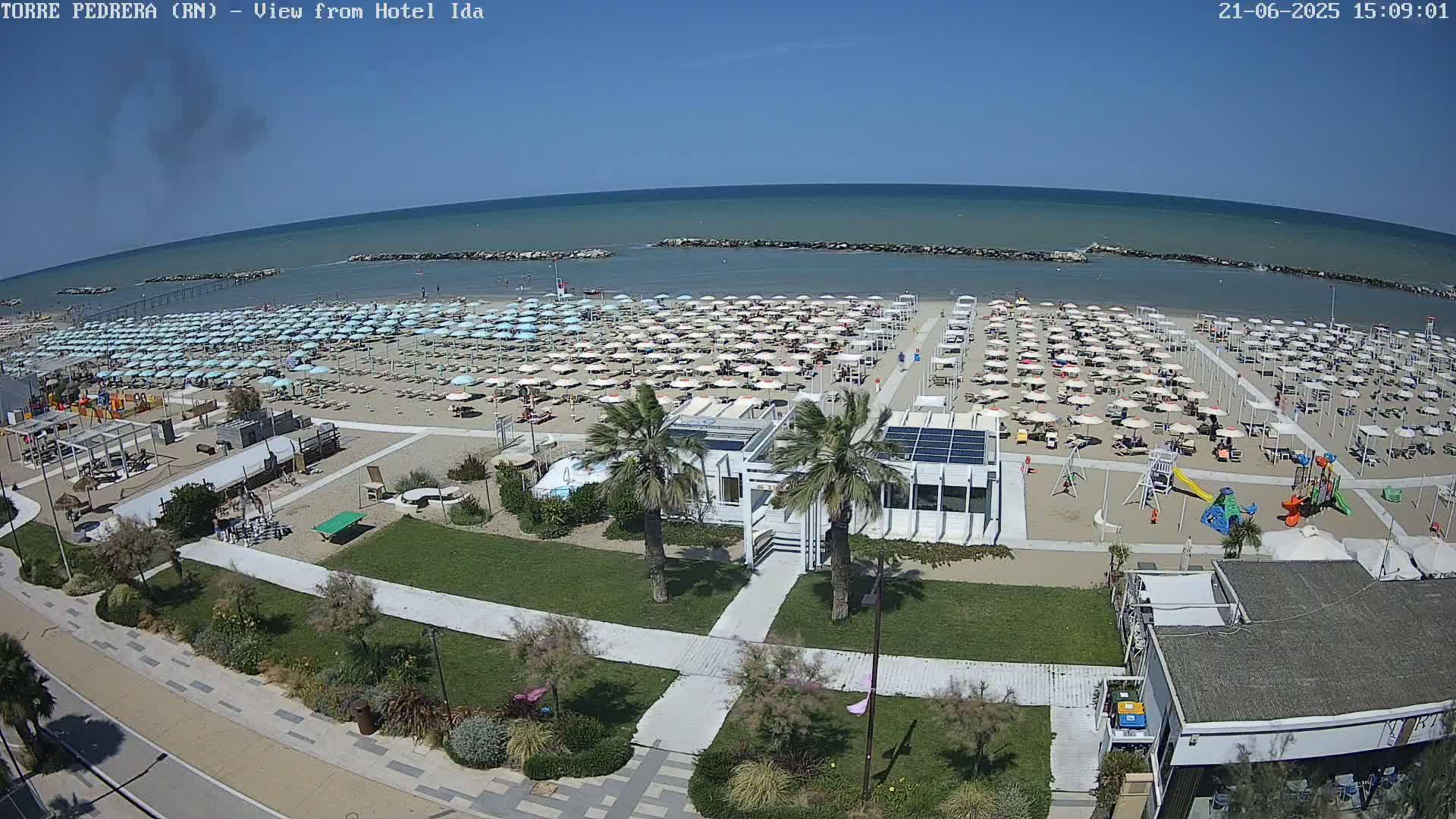 A sandy beach with numerous umbrellas and lounge chairs under a clear blue sky is shown, next to a grassy area with palm trees and a small building.