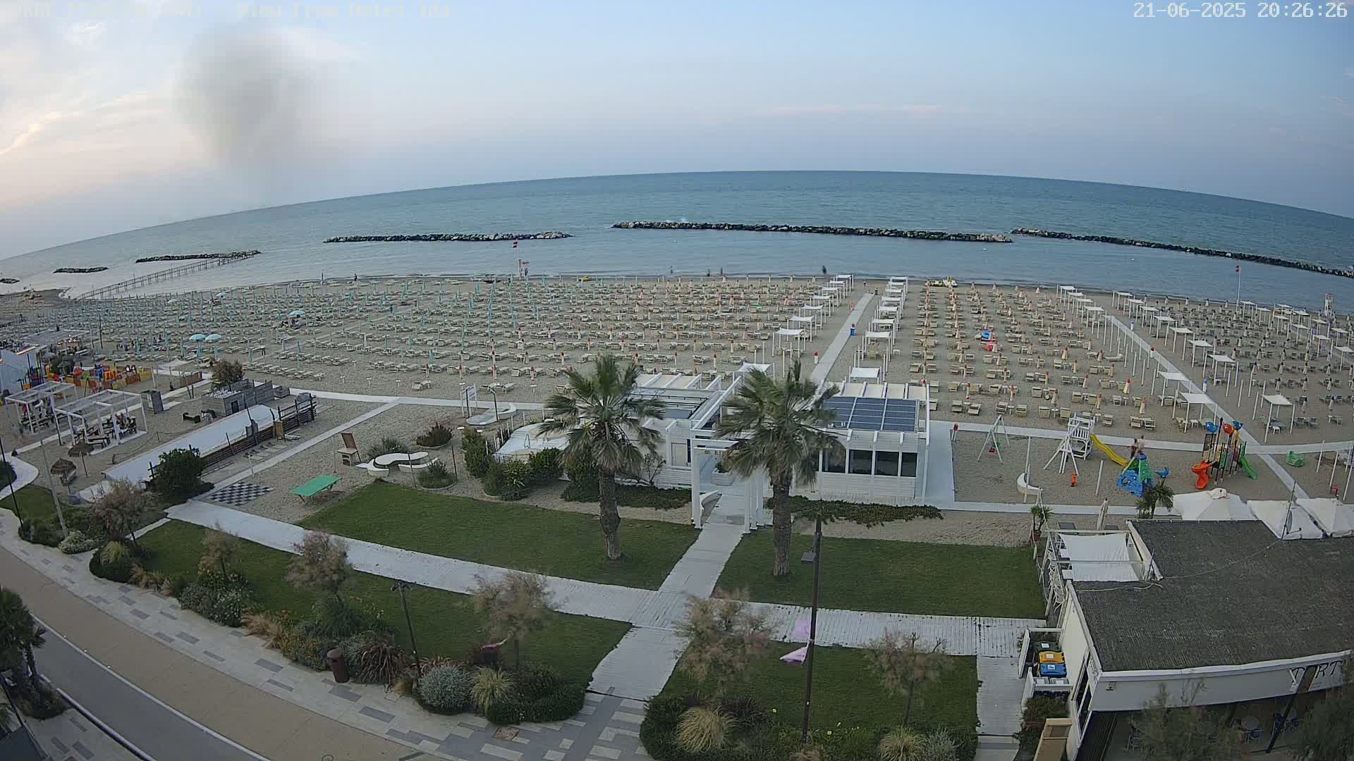 An expansive beach with rows of lounge chairs and umbrellas under a clear, twilight sky, features a beachfront building and a playground near a calm ocean.