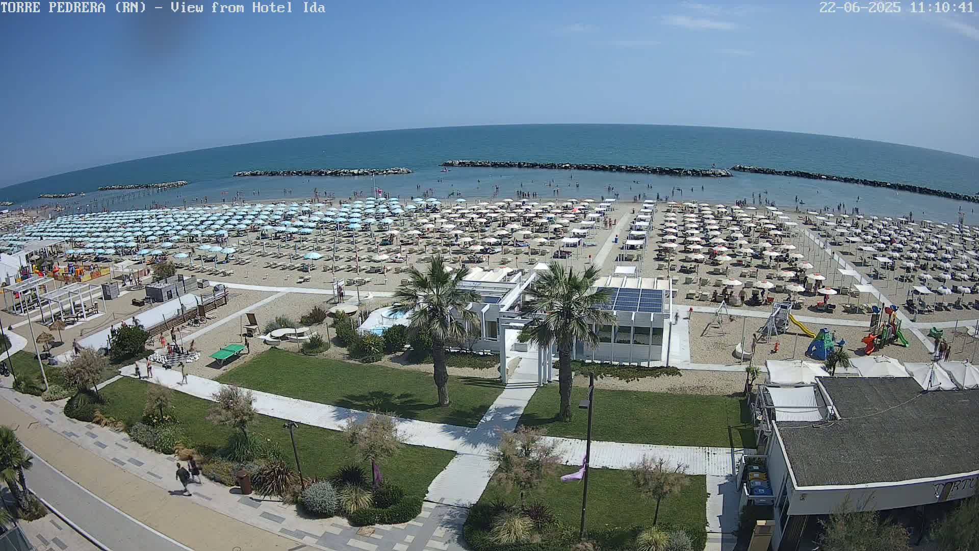 A sunny day at a crowded beach with numerous umbrellas and lounge chairs, a playground, and beachfront buildings.