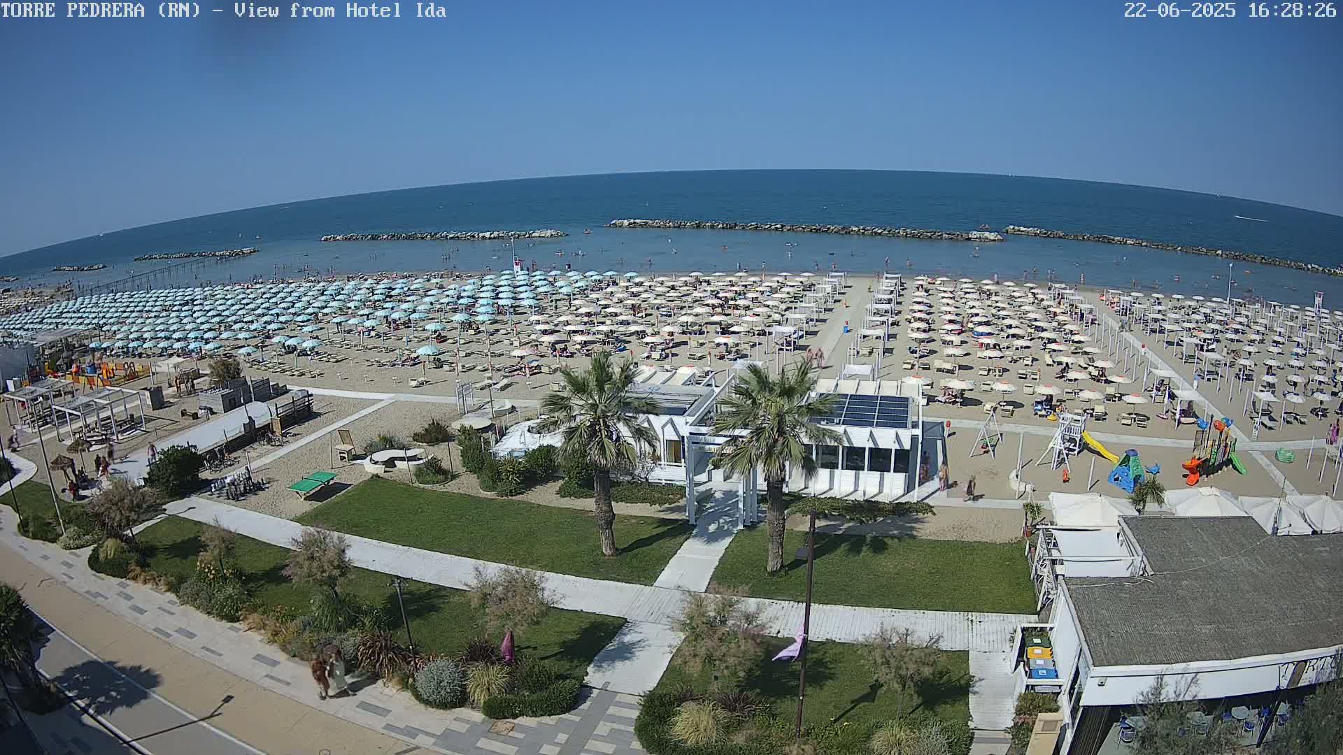 A crowded beach with numerous umbrellas and lounge chairs under a clear blue sky, adjacent to a grassy area with pathways, palm trees, and small buildings.