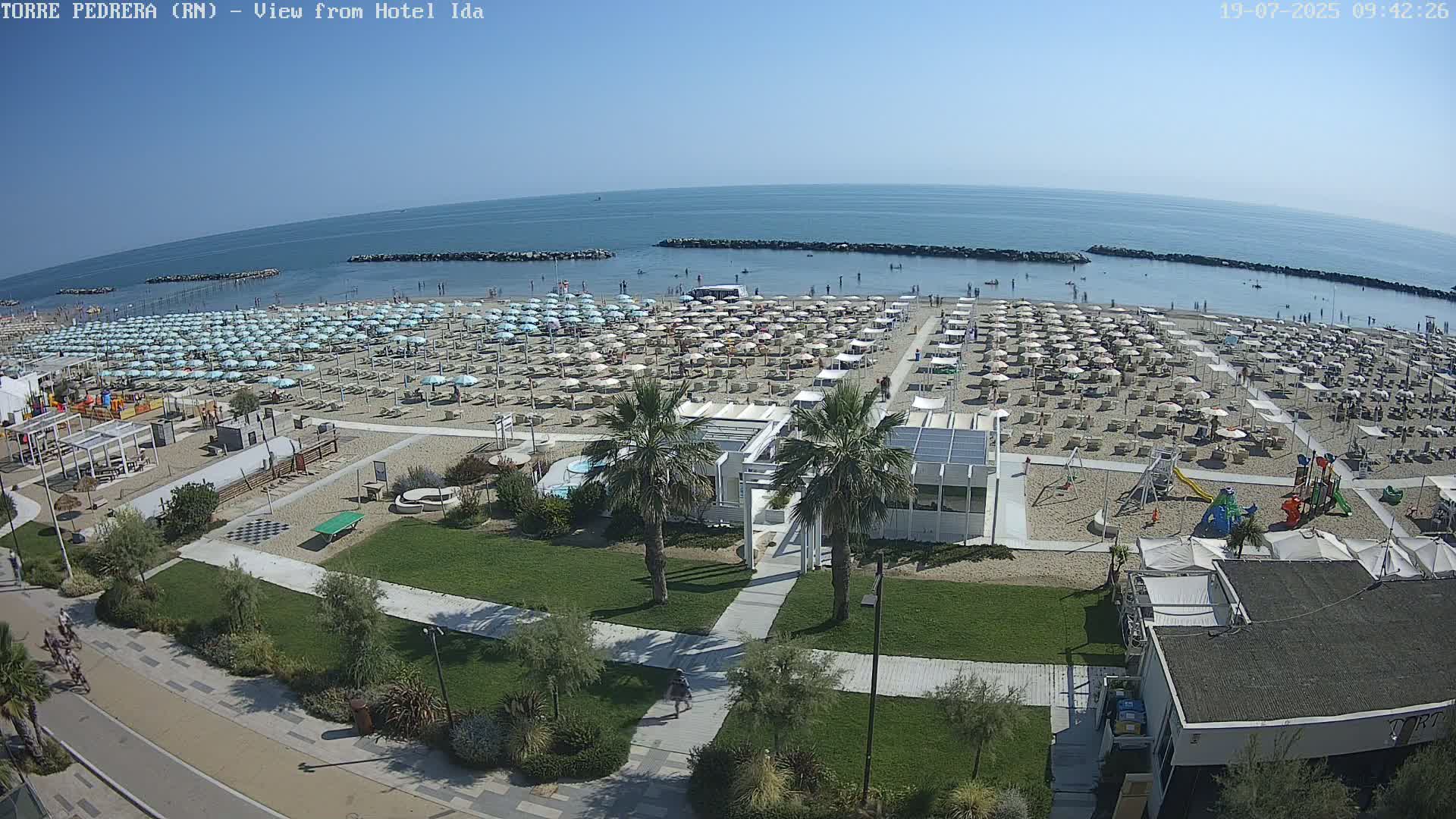A sandy beach with numerous umbrellas and beachgoers is visible from an elevated viewpoint, under sunny skies, with calm ocean waters in the background.