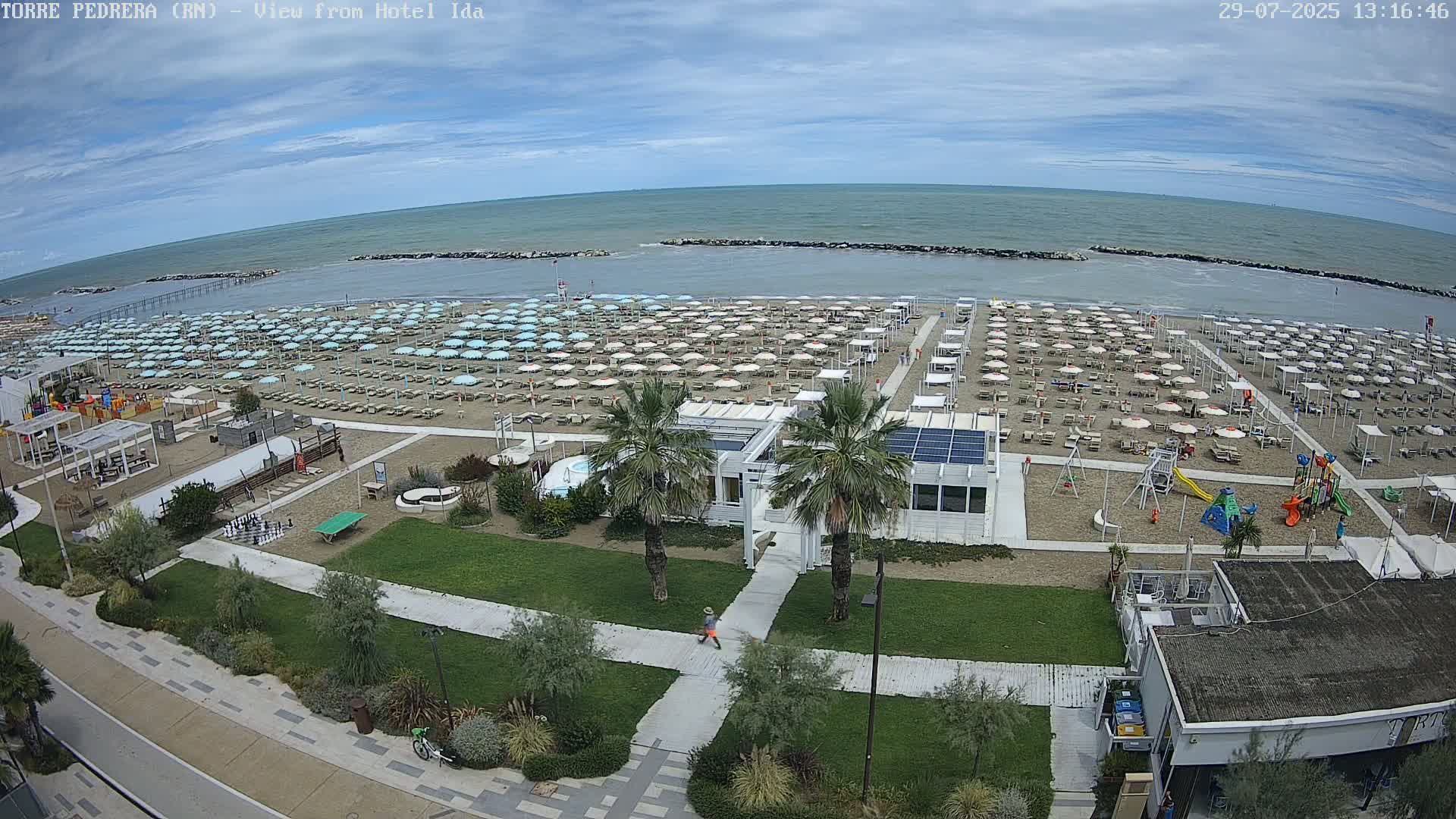 A sandy beach with numerous umbrellas and beachgoers is visible from an elevated viewpoint, under sunny skies, with calm ocean waters in the background.