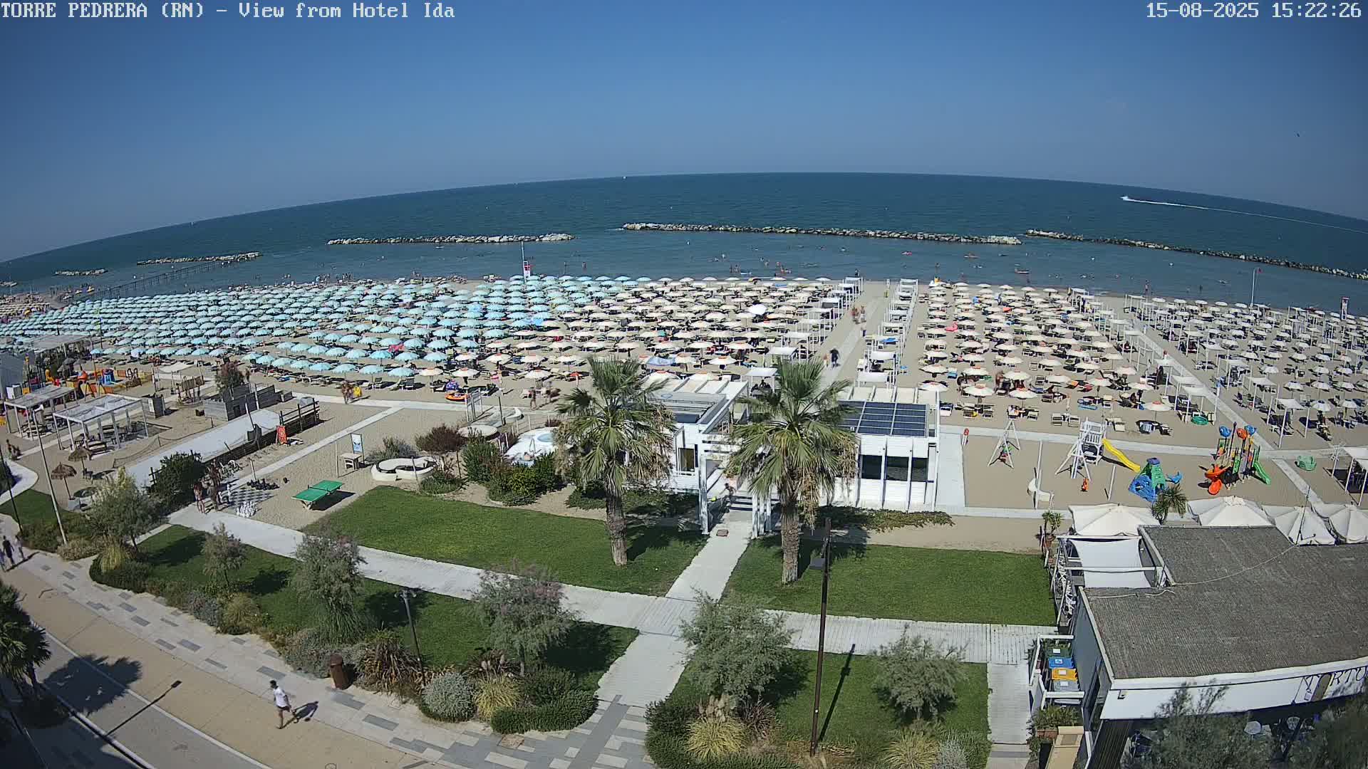 A beach with numerous umbrellas and sunbathers is shown, under a clear sunny sky, next to a grassy area with walkways, palm trees, and a playground.