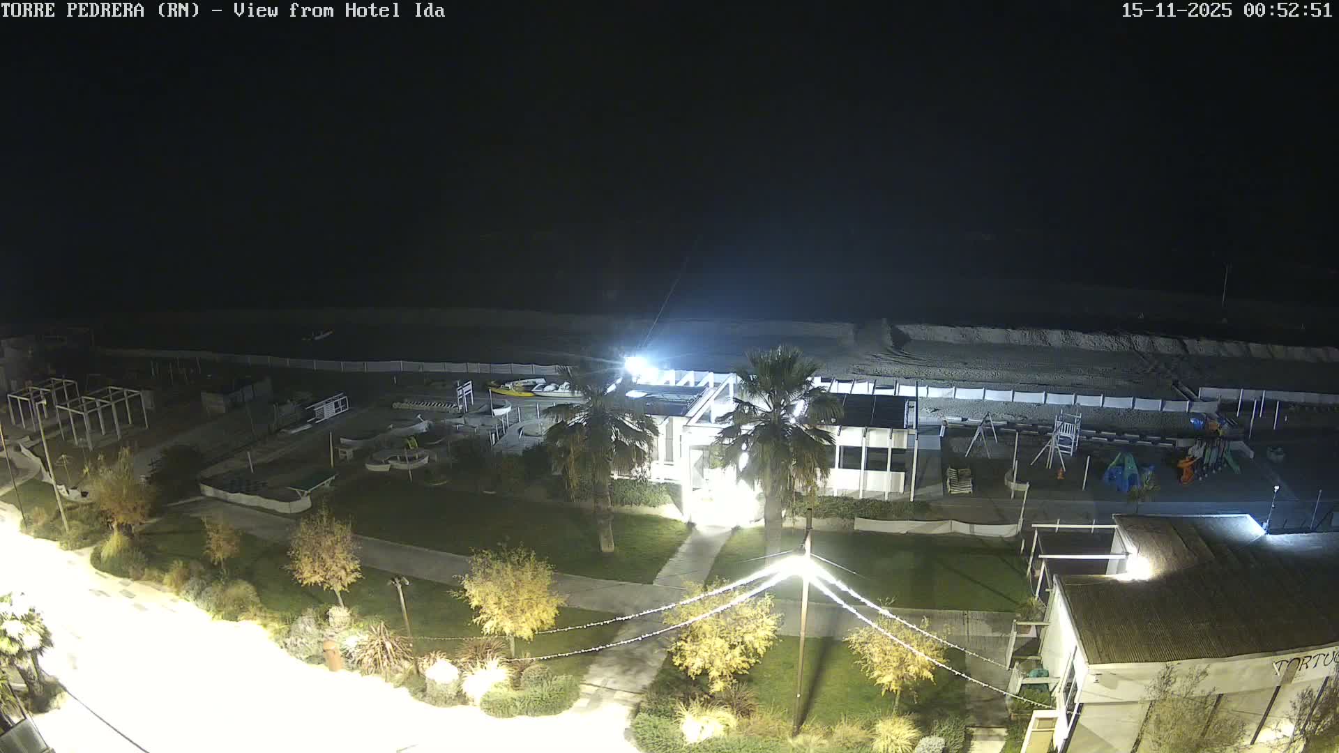The image shows an illuminated beach resort area at night with buildings, palm trees, pathways, and beach equipment on a sandy shore next to a dark, clear sea under a clear sky.