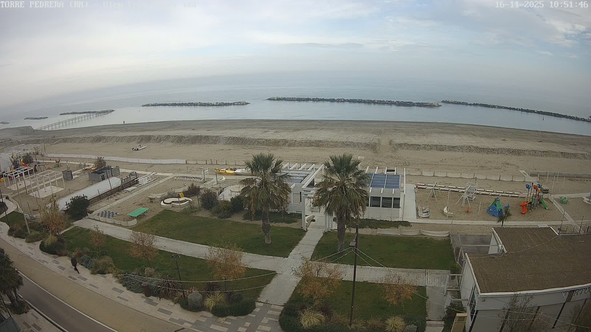 An overcast sky hangs over a calm sea with distant breakwaters and a pier, bordering a sandy beach featuring several beach clubs, a playground, green landscaped areas with palm trees, and a paved promenade.