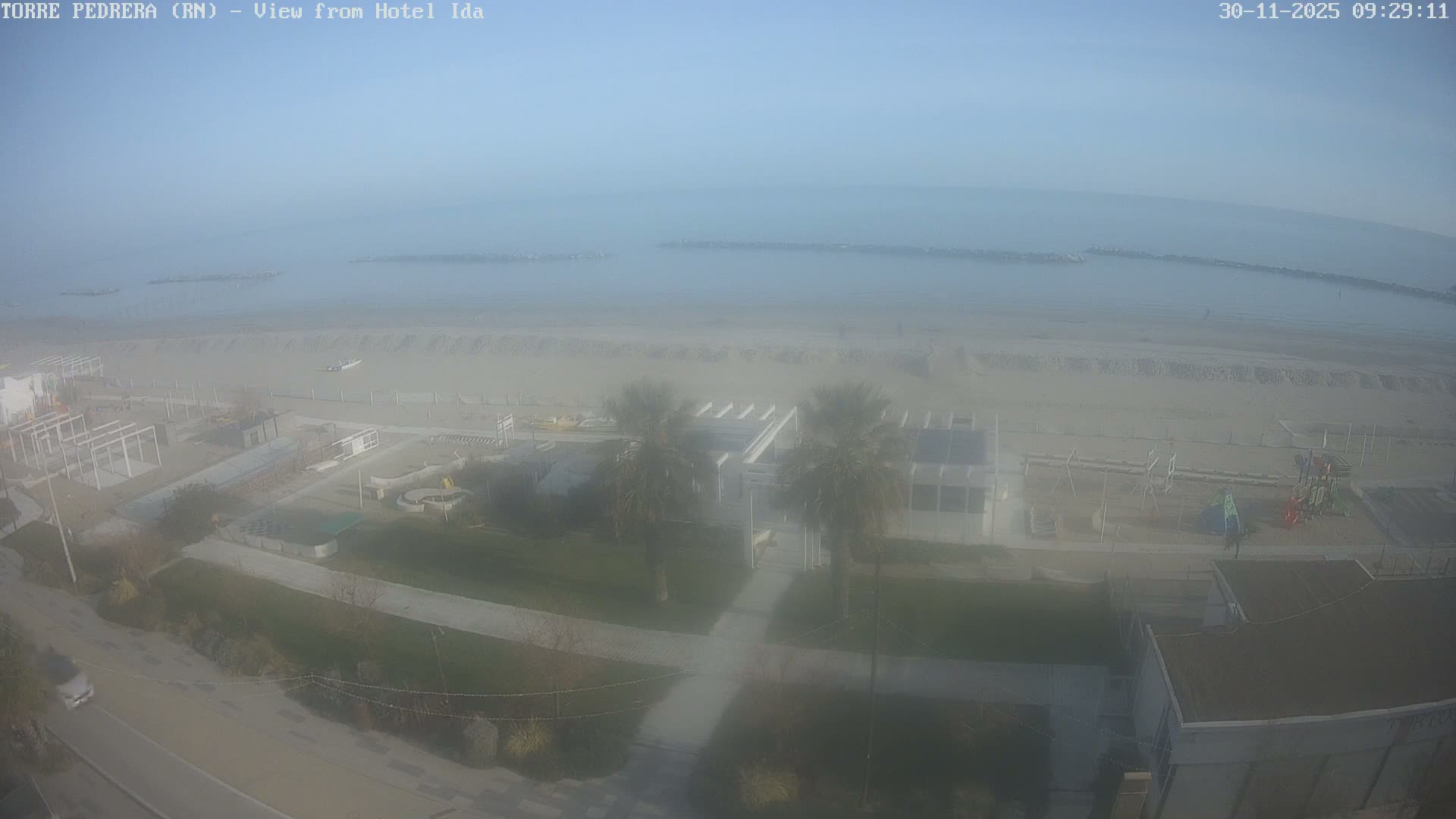The image displays a hazy daytime view of a wide sandy beach alongside a calm sea with visible breakwaters, bordered by various coastal buildings, palm trees, a playground, and a road with a car in the foreground.