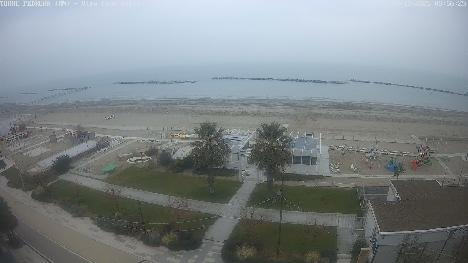 An overcast or hazy sky hangs over a wide, sandy beach featuring playgrounds, buildings, and palm trees, leading to a calm grey sea with distant breakwaters and a pier.