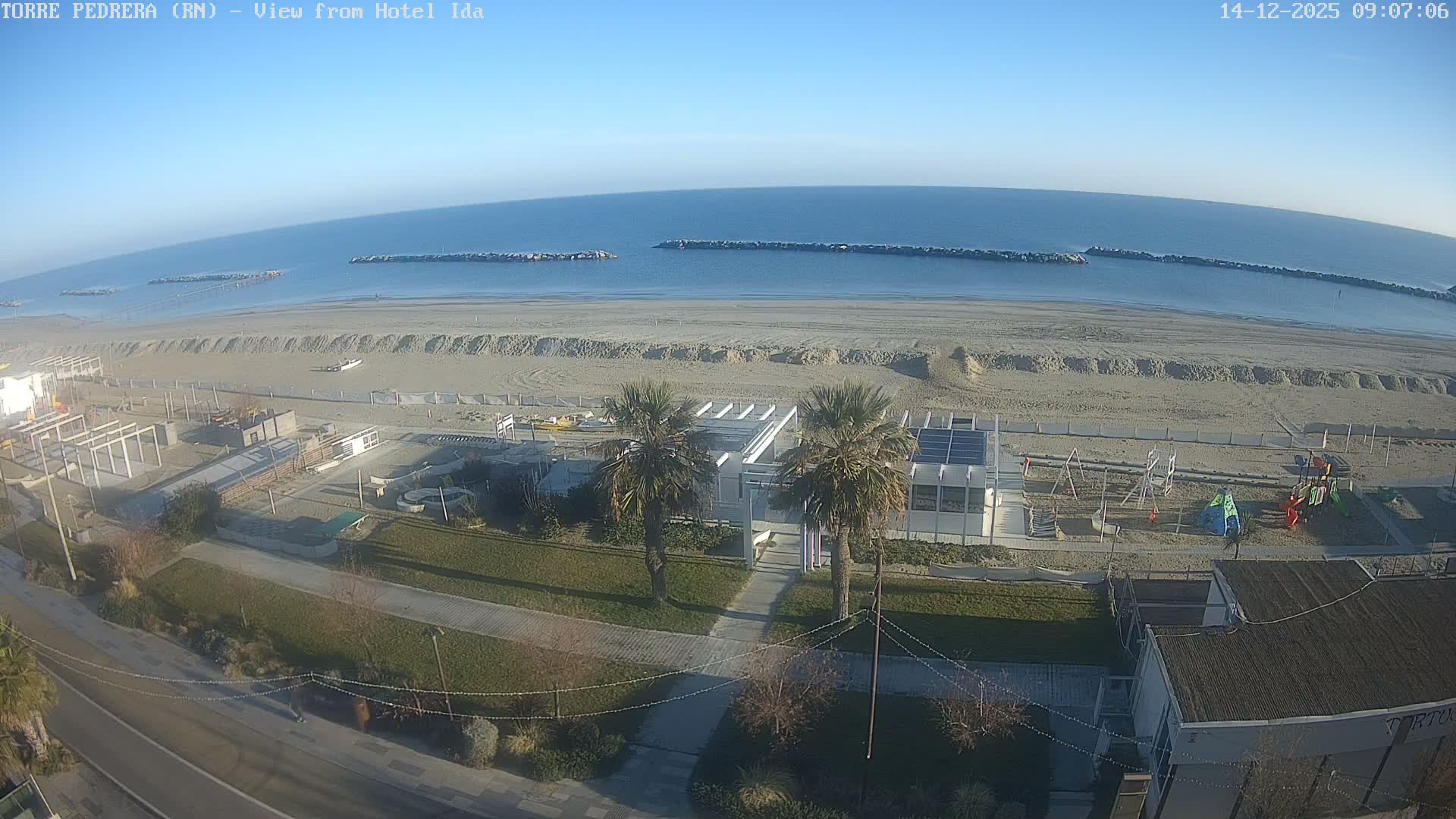 A wide sandy beach with scattered coastal buildings, palm trees, and a children's playground stretches towards the calm blue sea dotted with stone breakwaters, all under a clear, sunny blue sky.