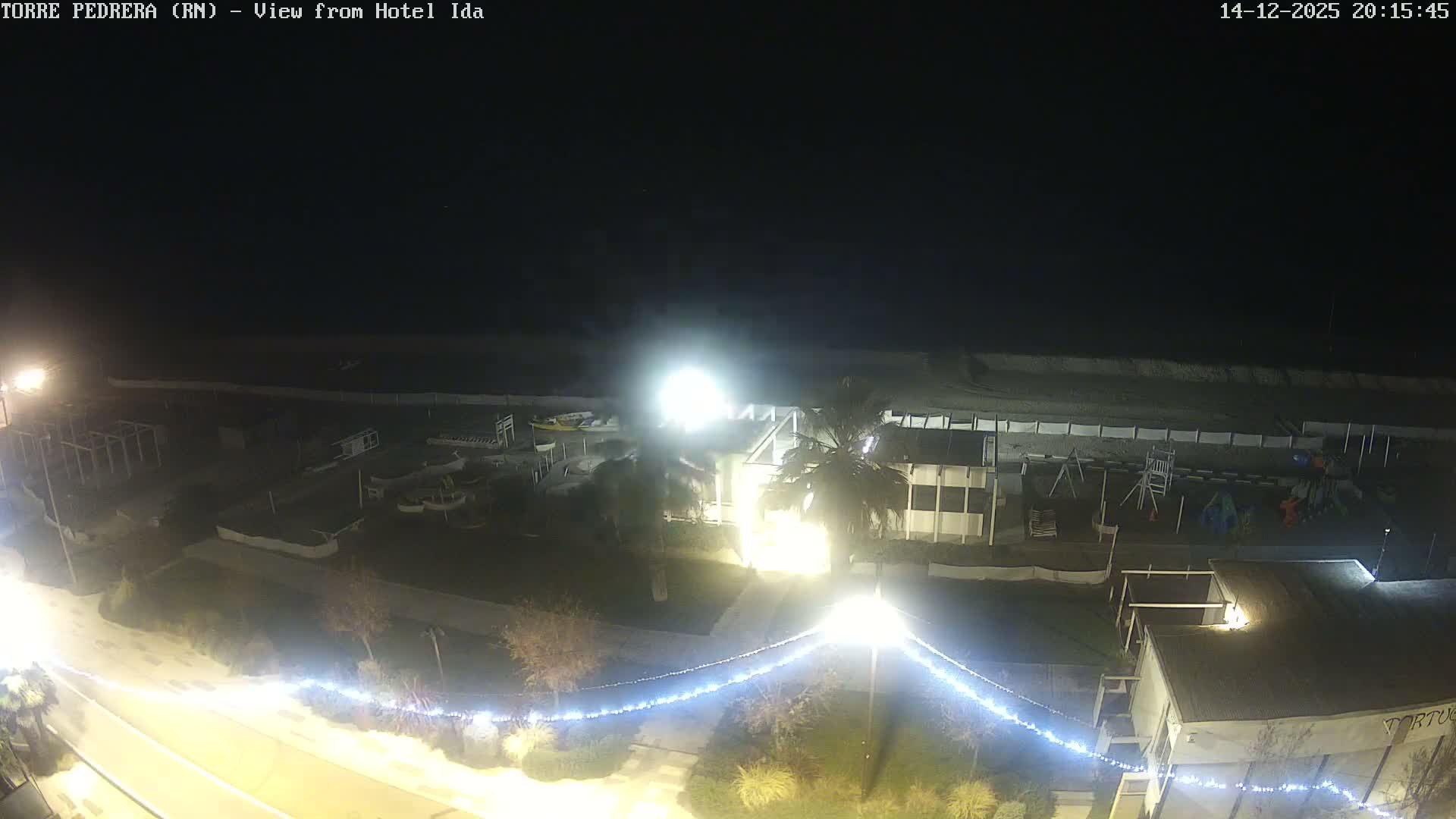 A wide sandy beach with scattered coastal buildings, palm trees, and a children's playground stretches towards the calm blue sea dotted with stone breakwaters, all under a clear, sunny blue sky.