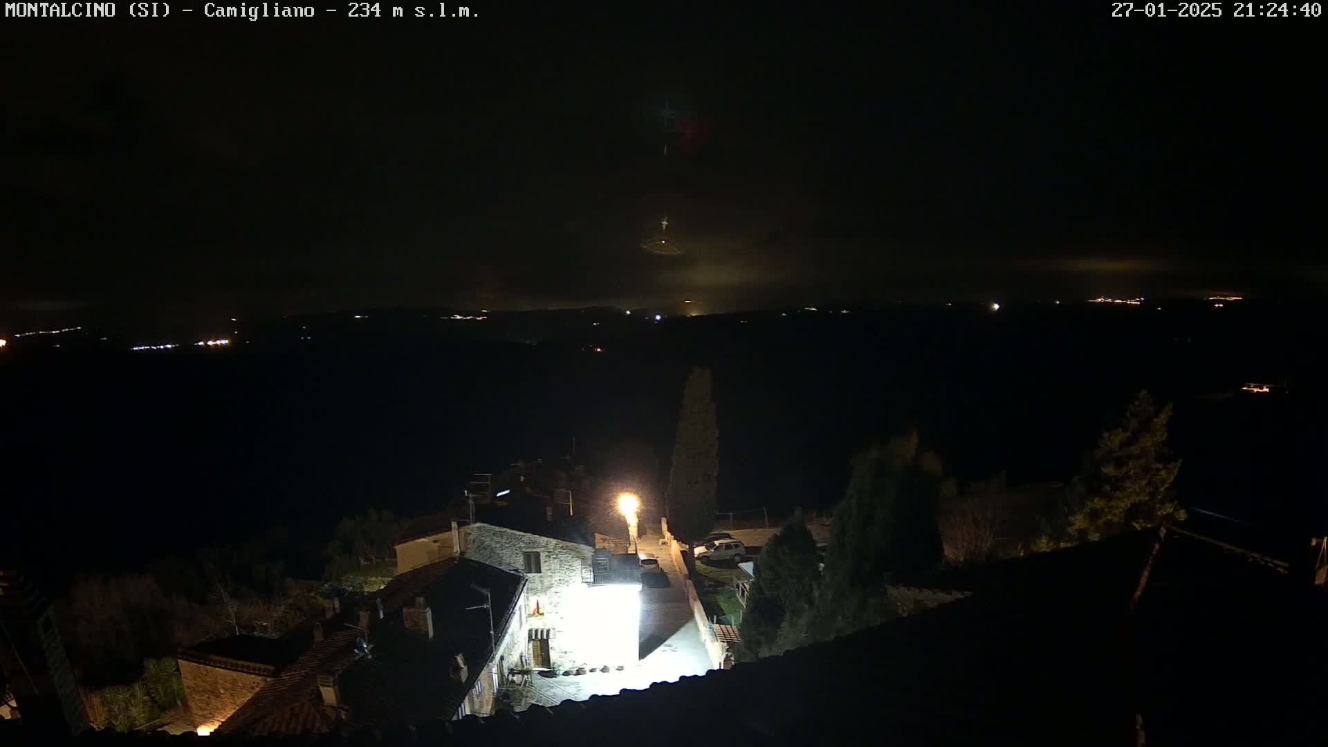 A nighttime, foggy view shows a village with dimly lit buildings and a hilltop in the distance.