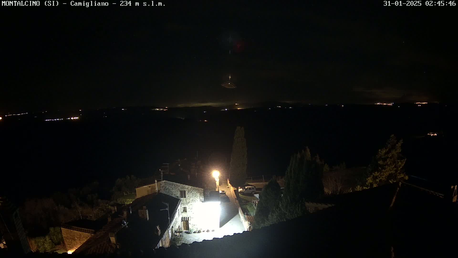 A nighttime aerial view shows a small village illuminated by a single bright light, overlooking a dark landscape dotted with distant lights under a mostly clear night sky.