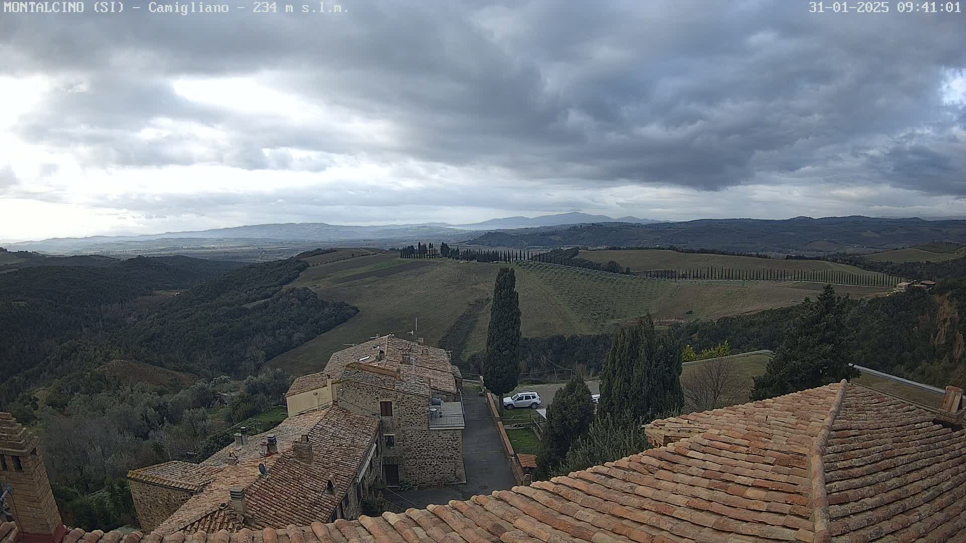 A view from a rooftop shows rolling hills, sparsely-treed farmland, and a small village under a cloudy sky.