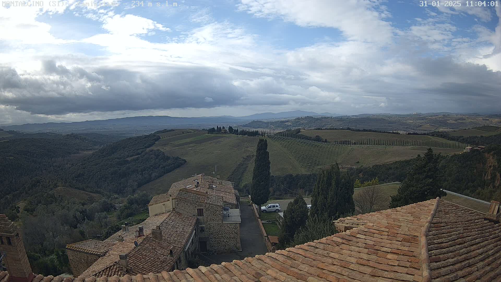 A partly cloudy sky overlooks a hilly landscape of olive groves and stone buildings with terracotta roofs.