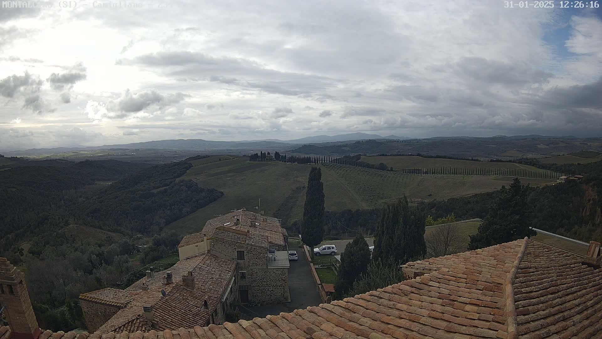 A view from a rooftop shows rolling hills under a cloudy sky, with stone buildings and cypress trees in the foreground.