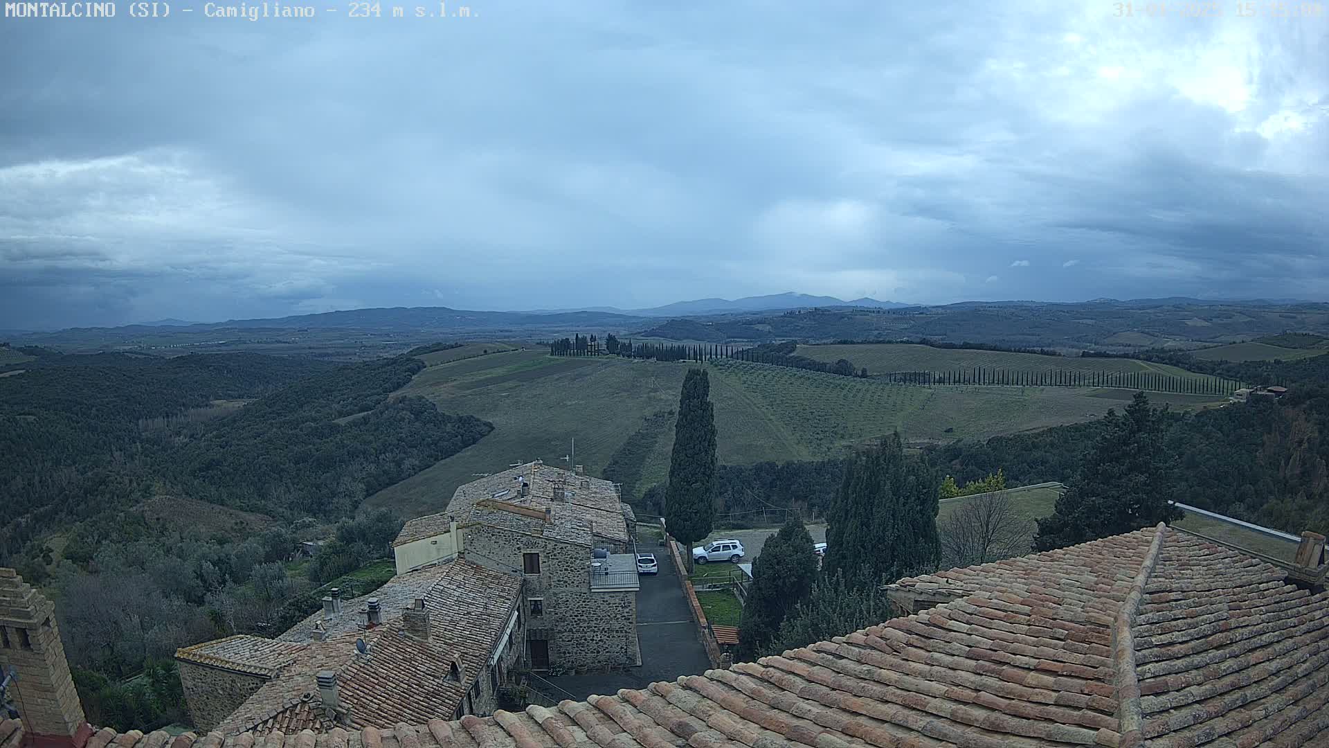 Overlooking a hilly landscape of olive groves and cypress trees under a cloudy sky, are the terracotta roofs of buildings in a small town.