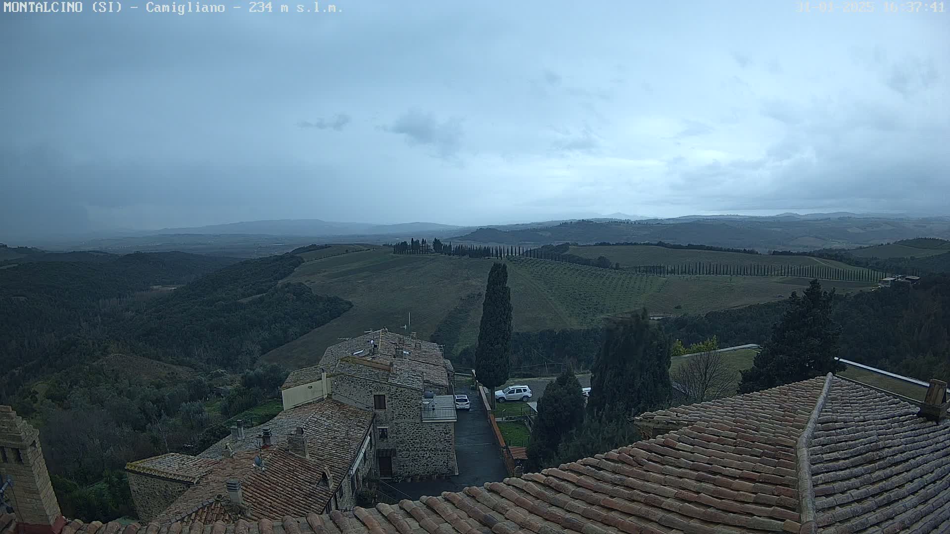 A view from a rooftop shows a cloudy sky over rolling hills dotted with trees and stone buildings, with a few cars visible in a small parking area.