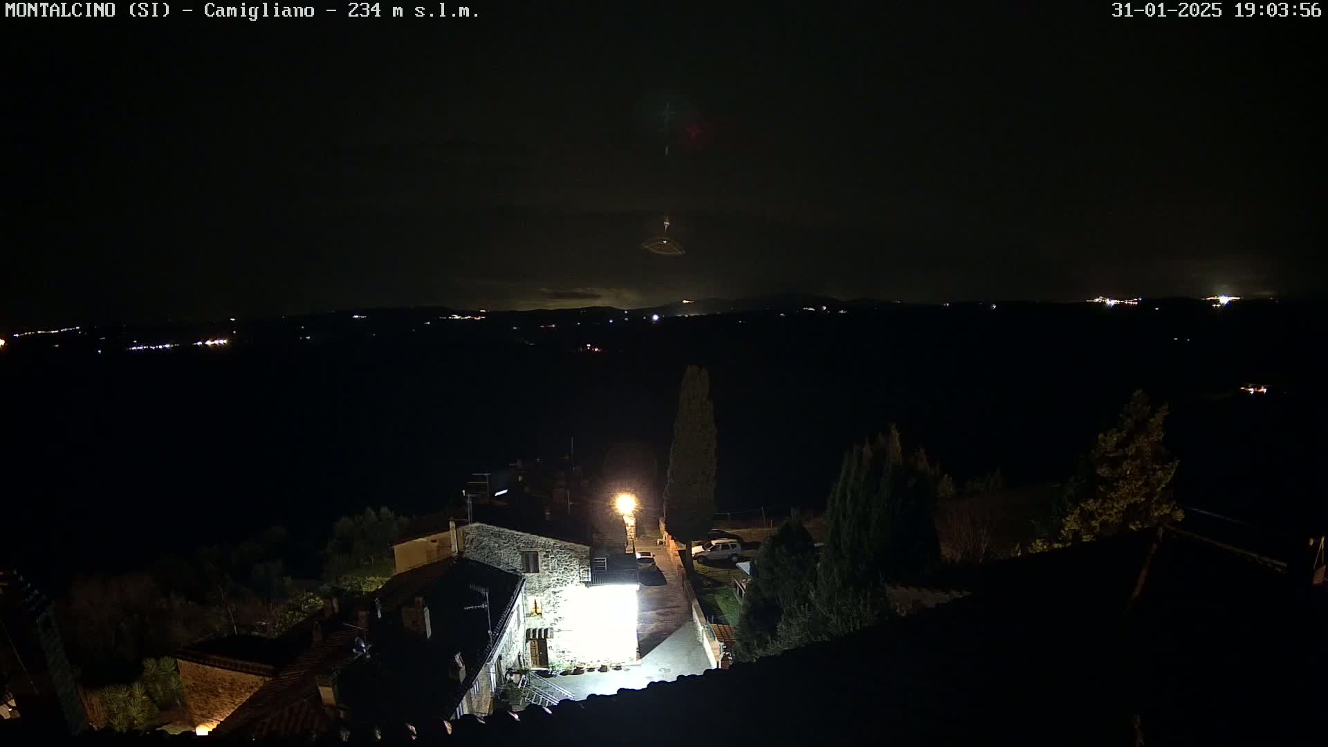A nighttime, high-angle view of a small village illuminated by scattered lights, with a distant, dark landscape showing faint lights and a bright object possibly in the night sky.