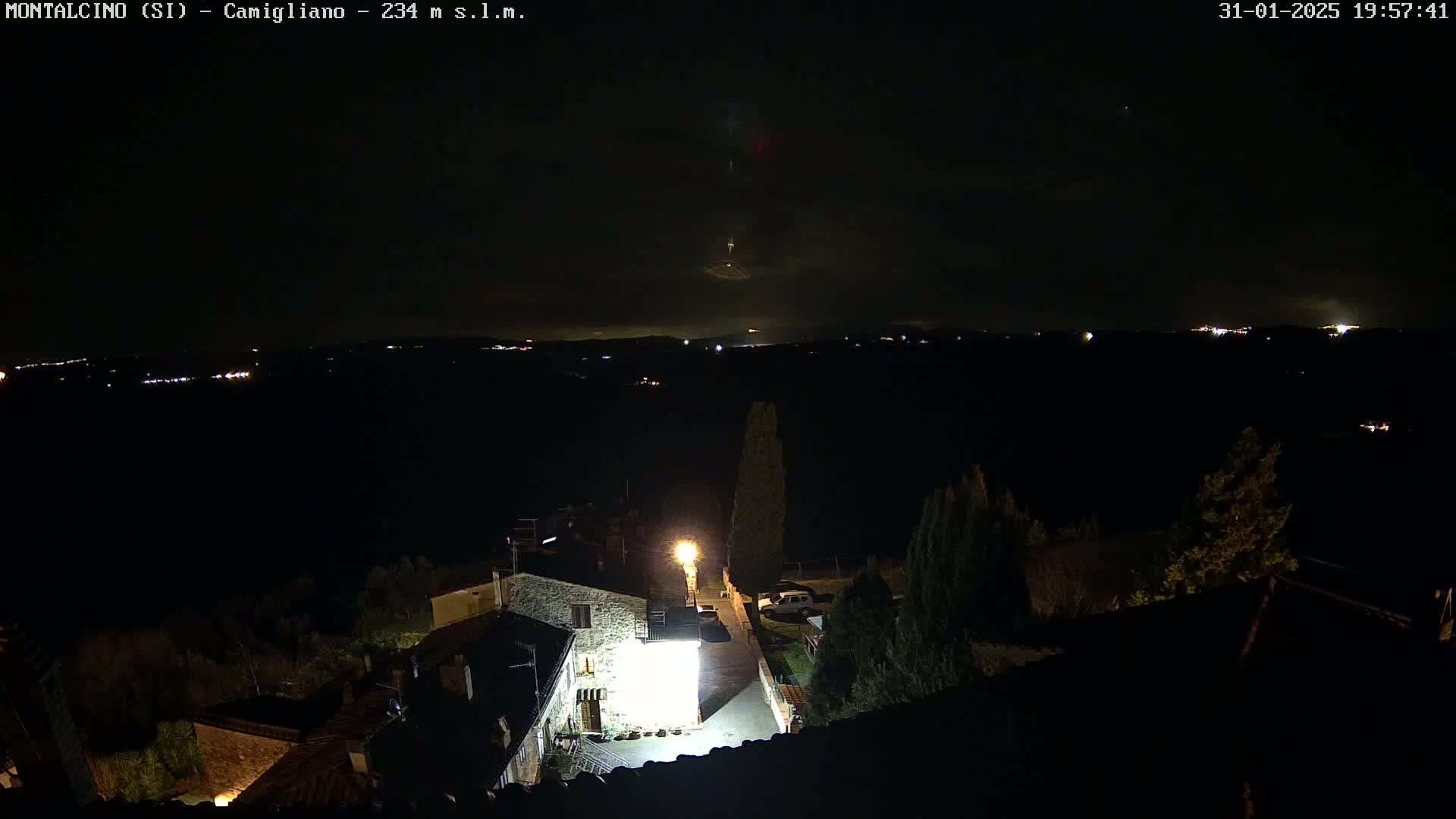 A nighttime, high-angle view shows a small village illuminated by lights, with a distant, faintly lit horizon under a dark, cloudy sky.
