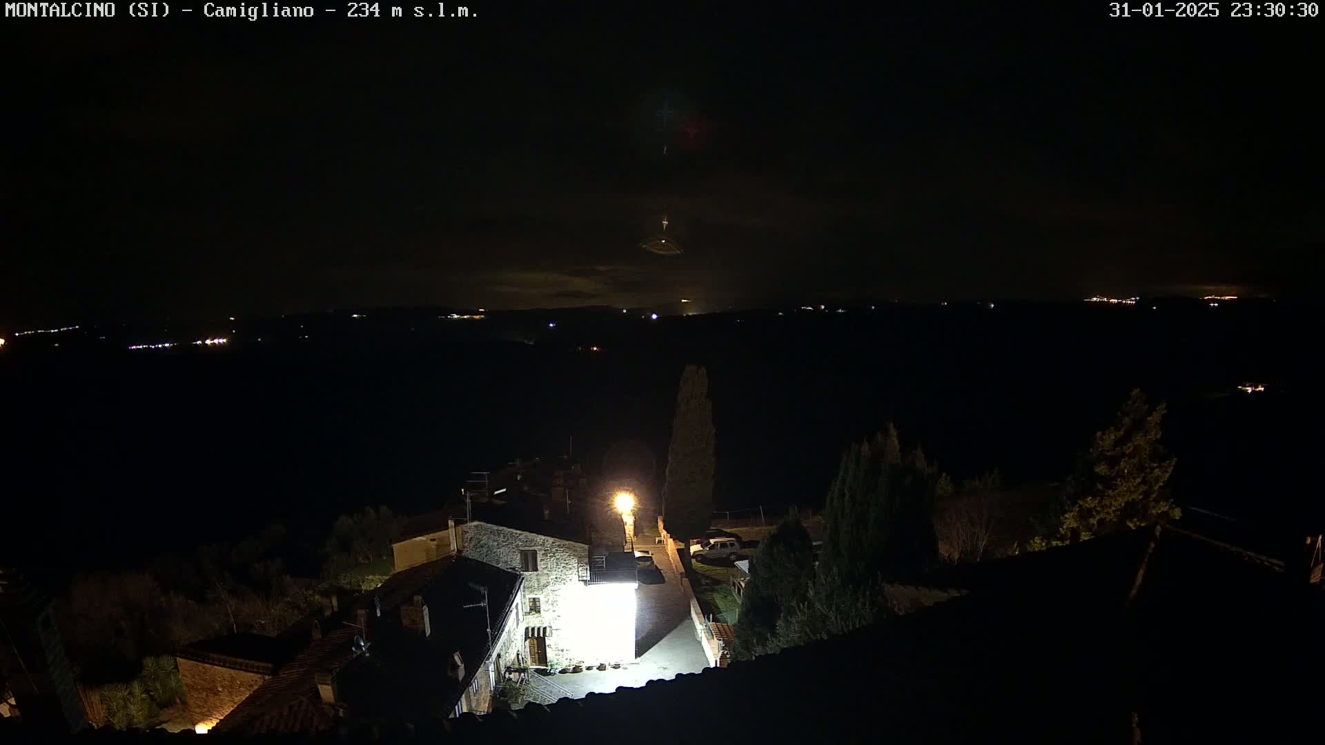 A nighttime view of a small village illuminated by a single bright light, overlooking a dark landscape with distant lights under a mostly clear sky.