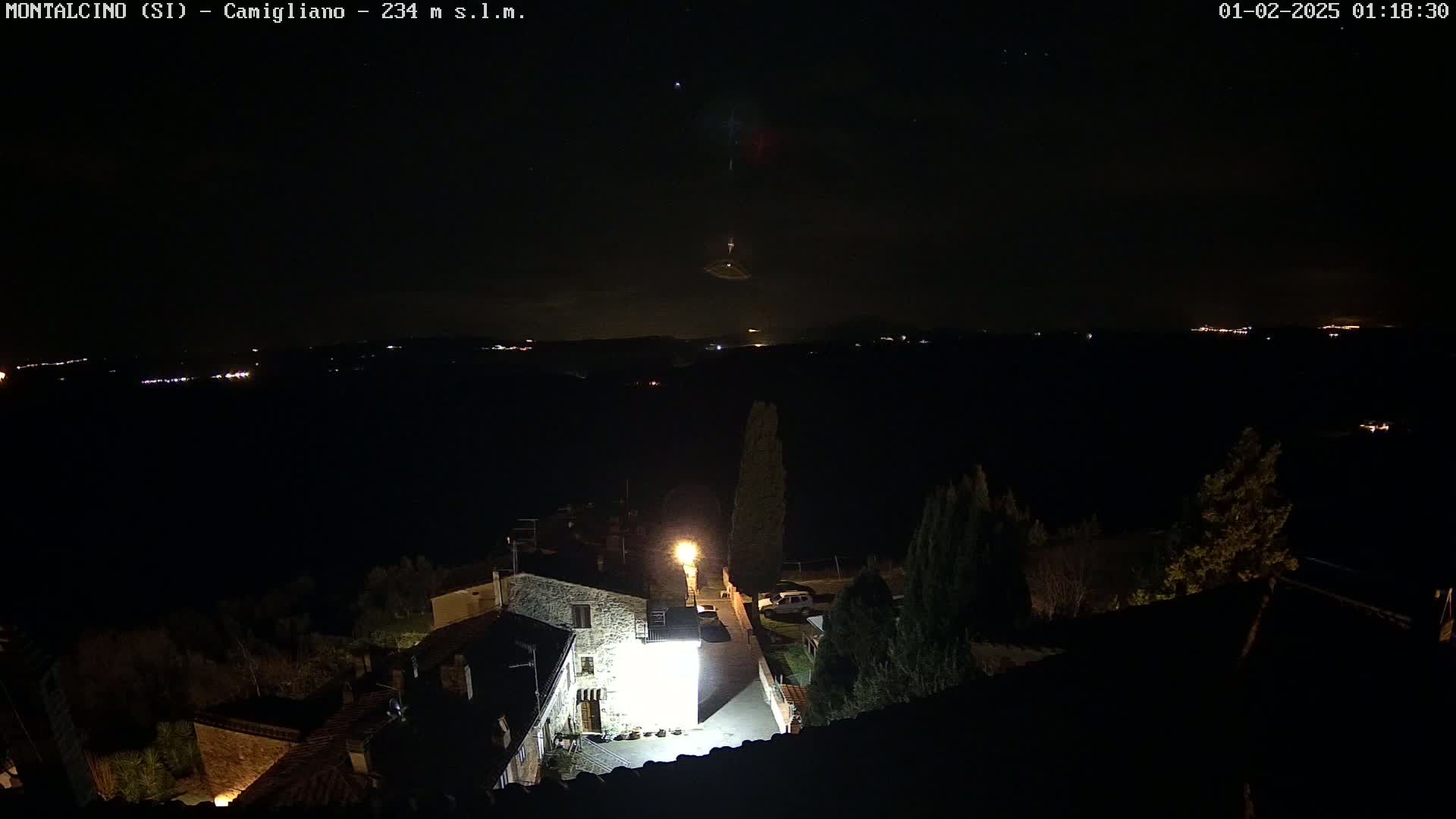 A nighttime, high-angle view of a small village illuminated by a bright light, overlooking a dark landscape dotted with distant lights under a clear, starry sky.